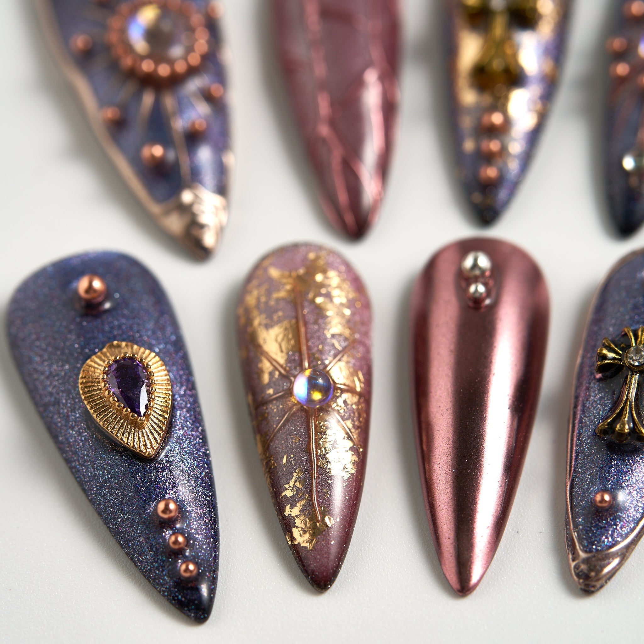 Macro of Violet Eclipse stiletto nails with metallic purple and gold details, rhinestones, and textured finishes on a white background