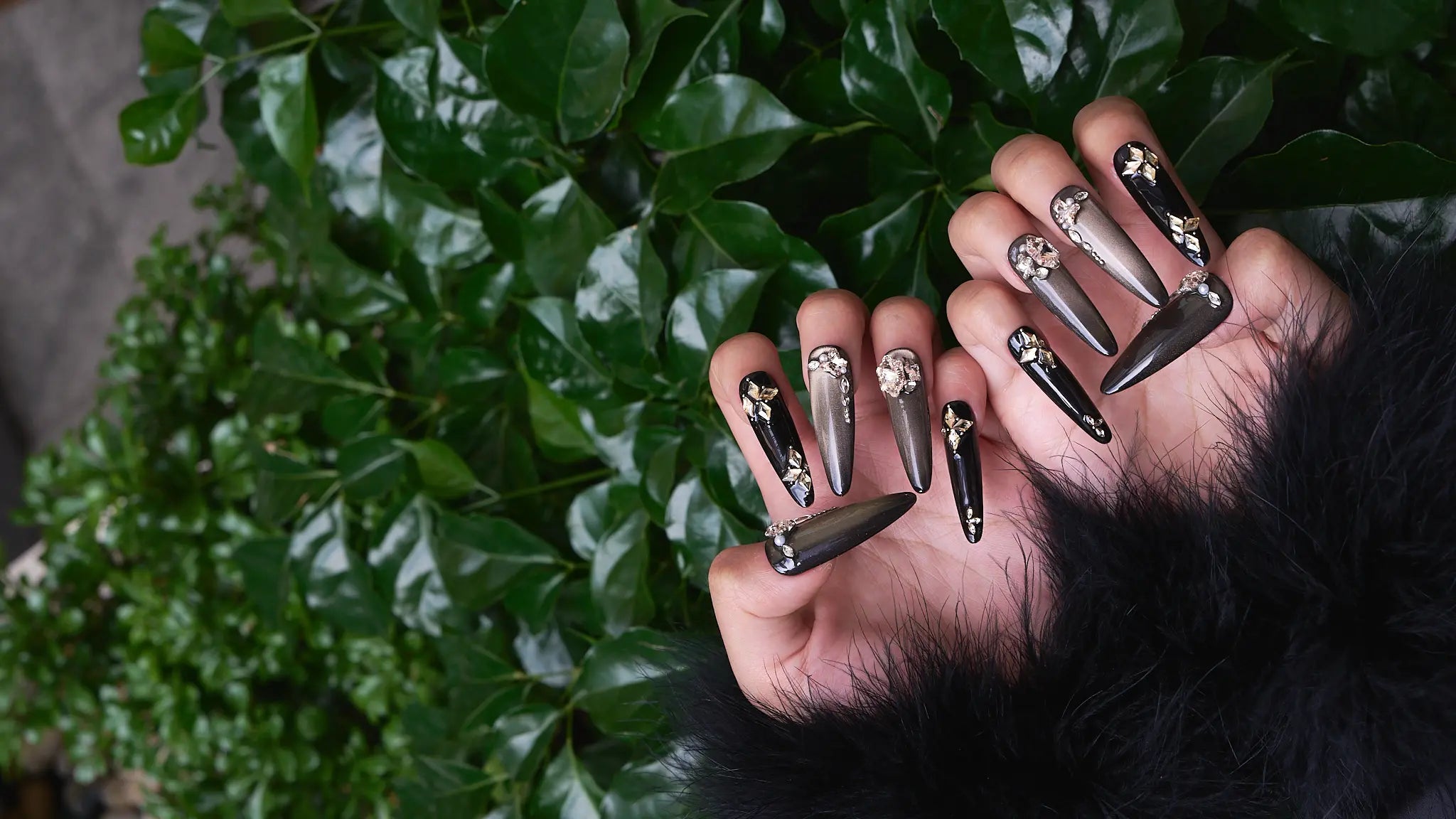 long stiletto press-on nails in metallic black with gold and crystal accents, shown against green leaves background