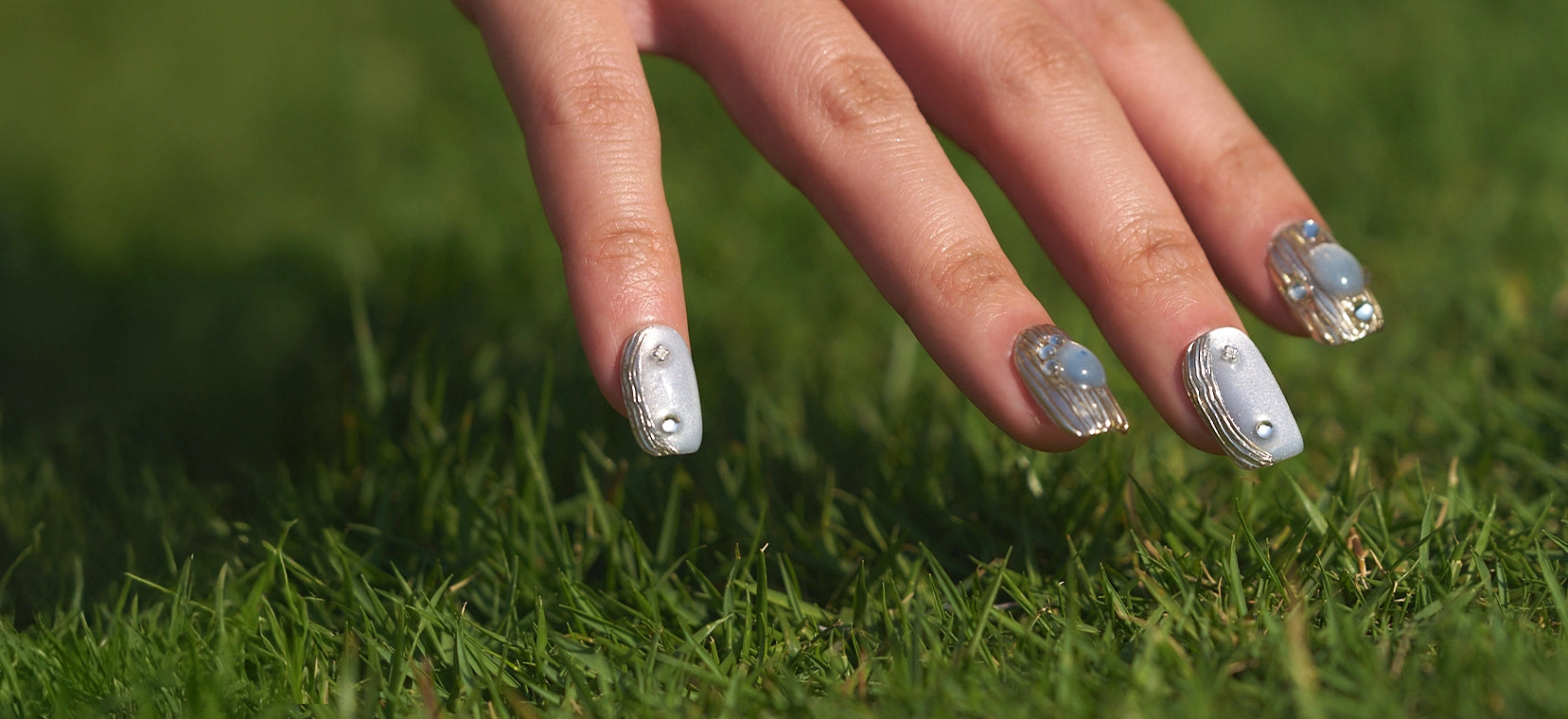 Hand wearing MiuNail “Mist Orbit” silver and blue press-on nails with rhinestone accents, photographed outdoors on green grass, featured in the About page hero banner.