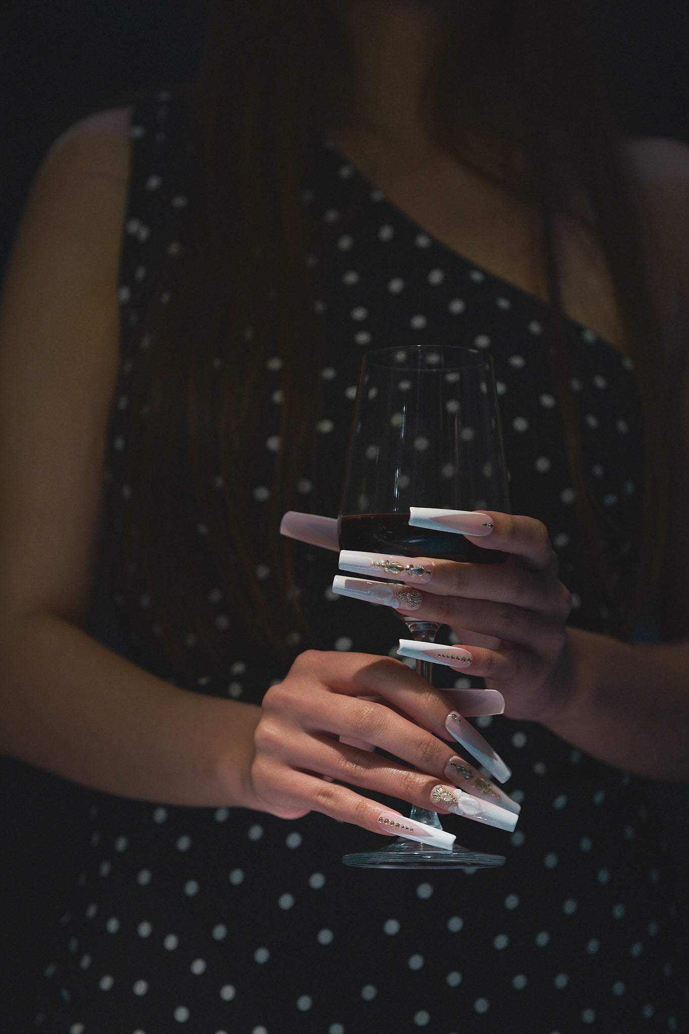 long square nails in blush pink and white with gold details and 3D bow holding a wine glass against a dark background in evening wear