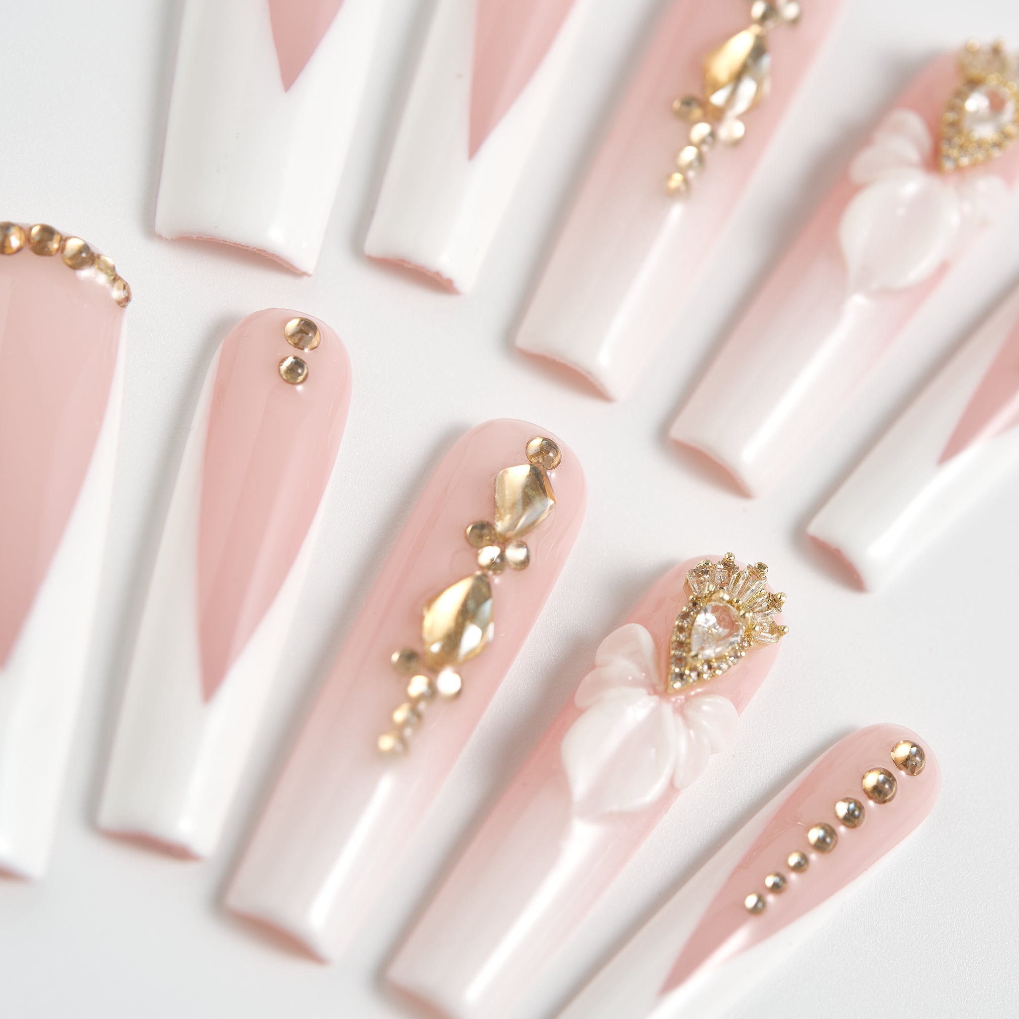 close-up of long square nails in blush pink and white with gold rhinestones and 3D bow details on a white background