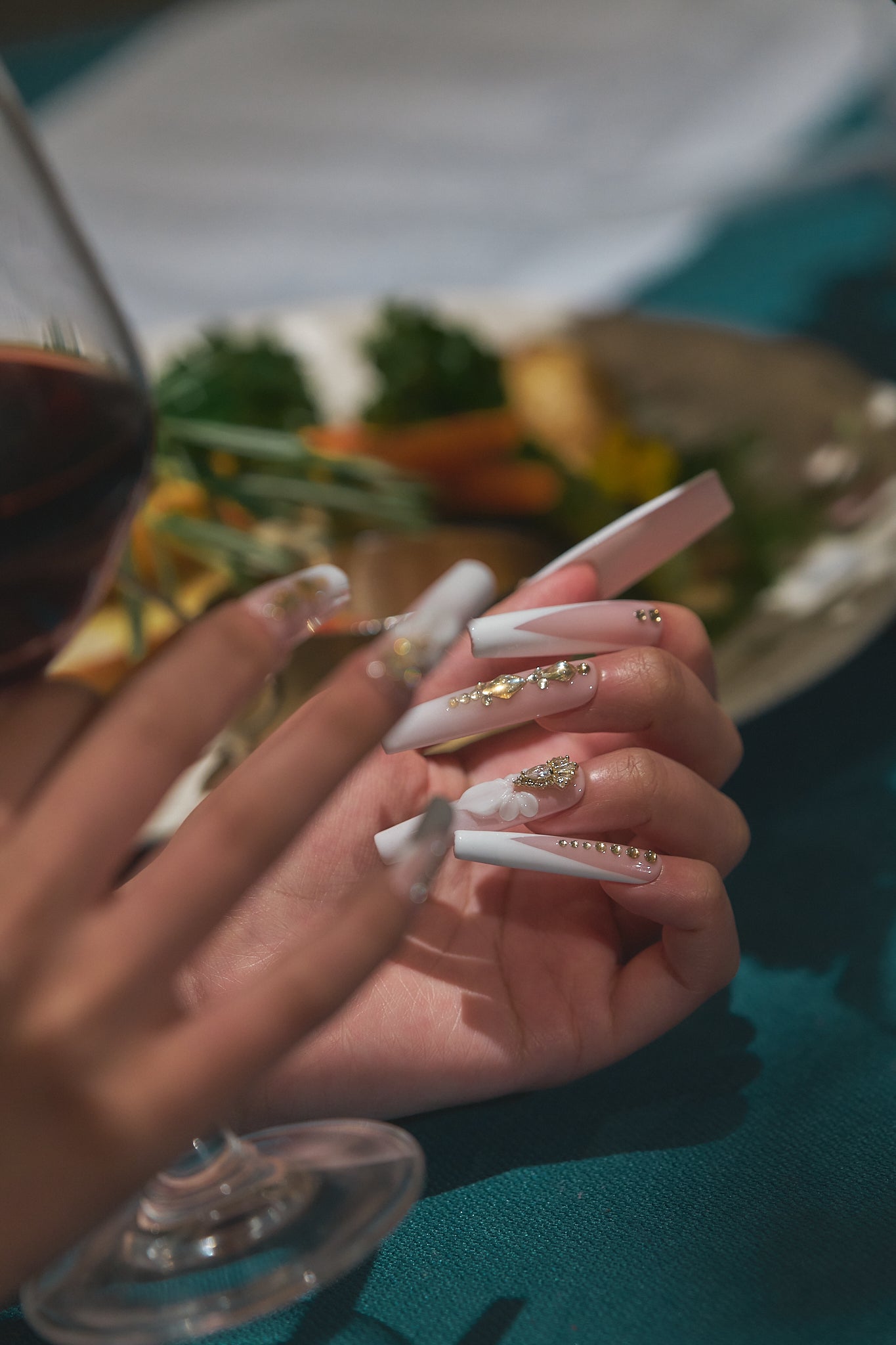 long square nails in blush pink and white with gold details and 3D bow holding a red wine glass over a blue table with blurred food in background
