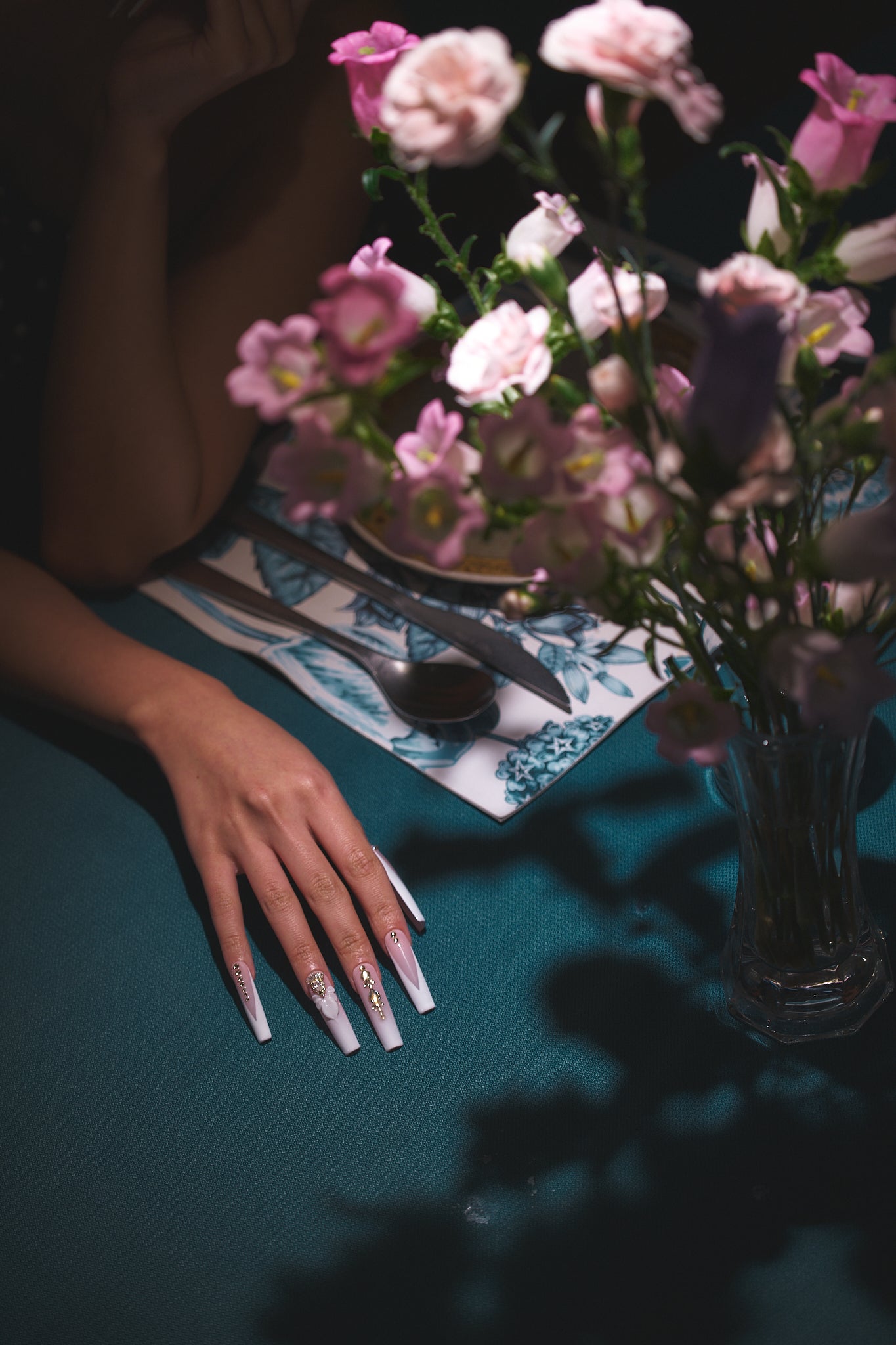 long square nails in blush pink and white with gold embellishments resting on a teal table beside cutlery and a vase of pink flowers