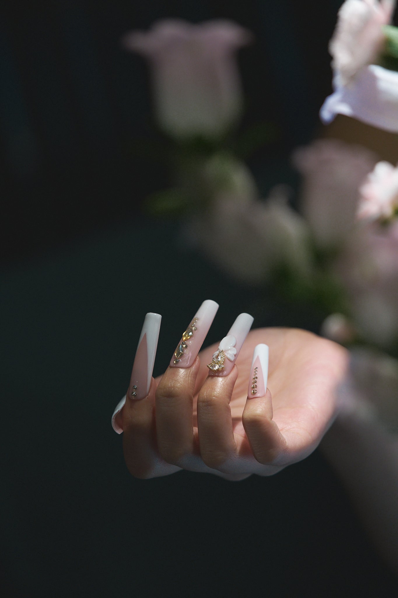 close-up of long square nails in blush pink and white with gold embellishments and 3D bow shown against a blurred floral background