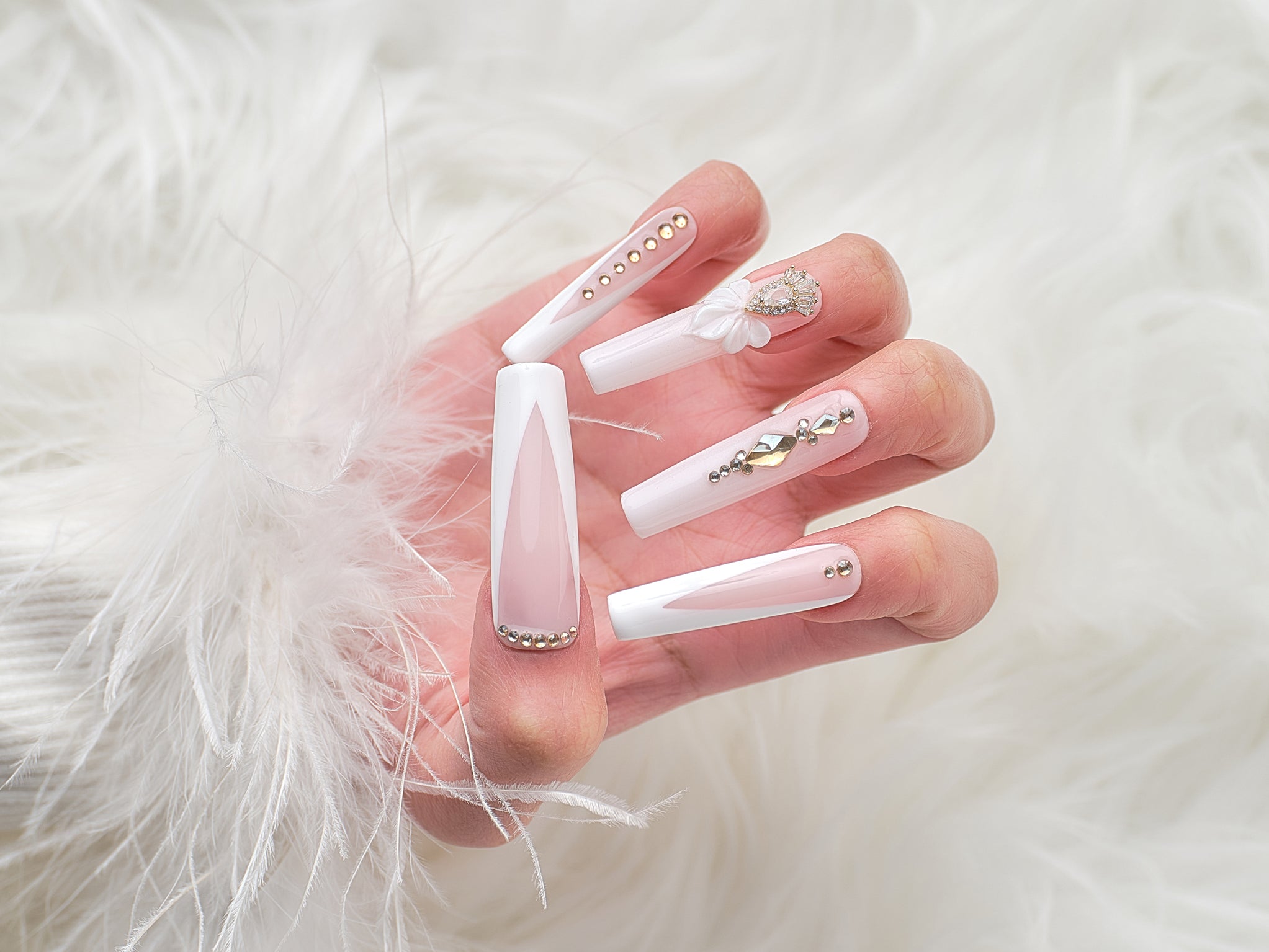 long square nails in blush pink and white with gold gems and 3D bow shown on a hand against white faux fur background