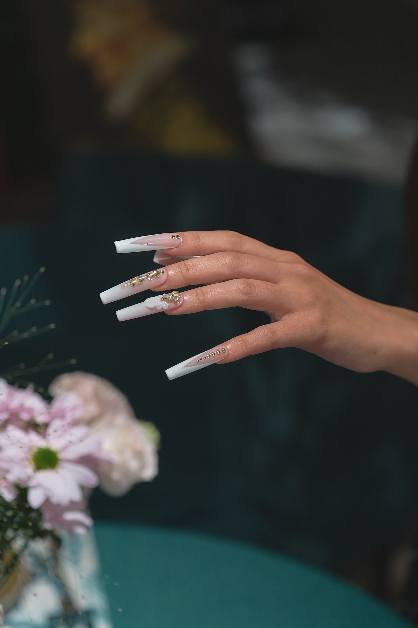 long square nails in blush pink and white with gold rhinestones and 3D bow shown on an extended hand near pink flowers on a blurred teal background