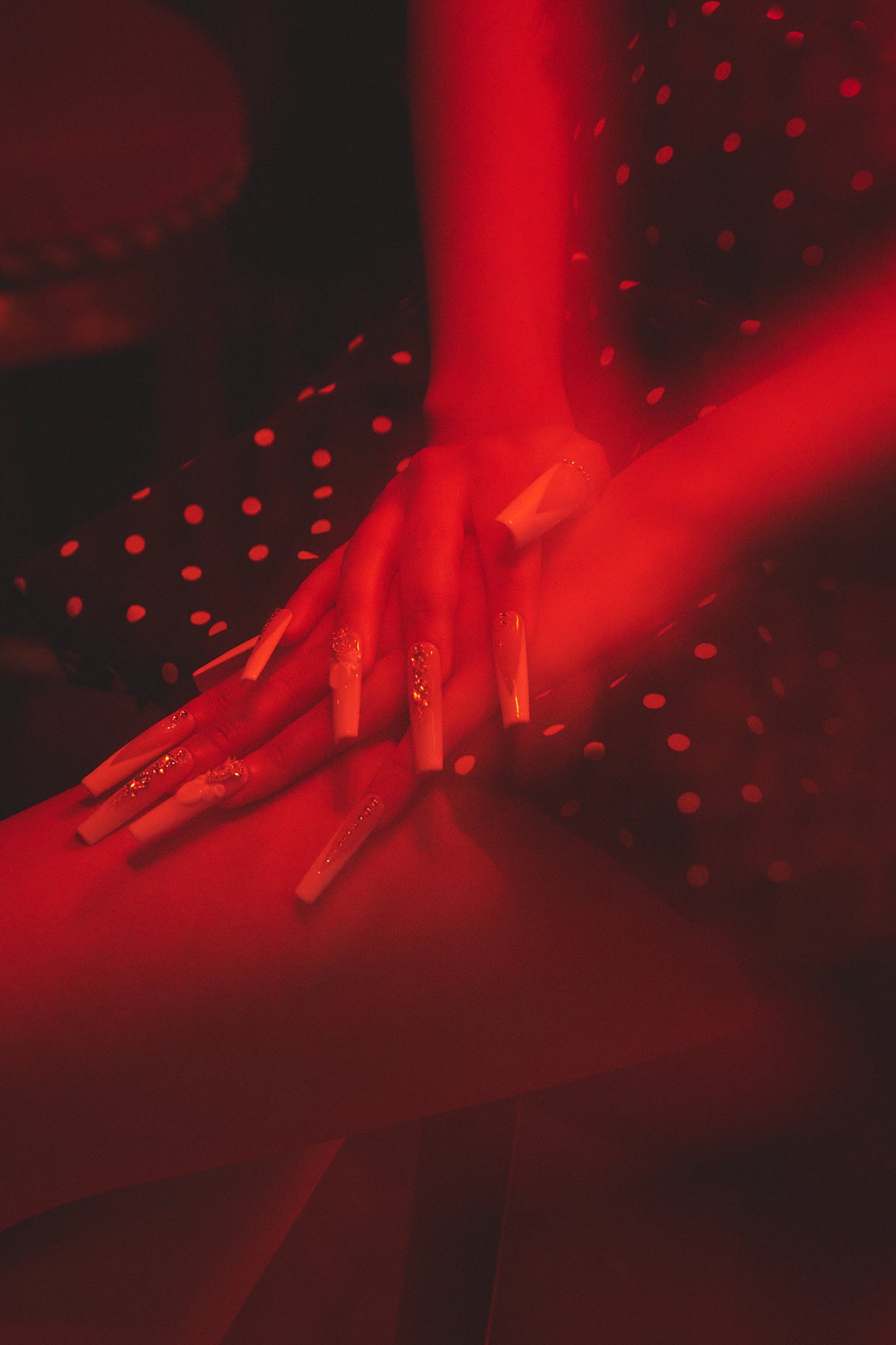 close-up of hands with blush pink long square nails resting on lap under red lighting in polka dot dress