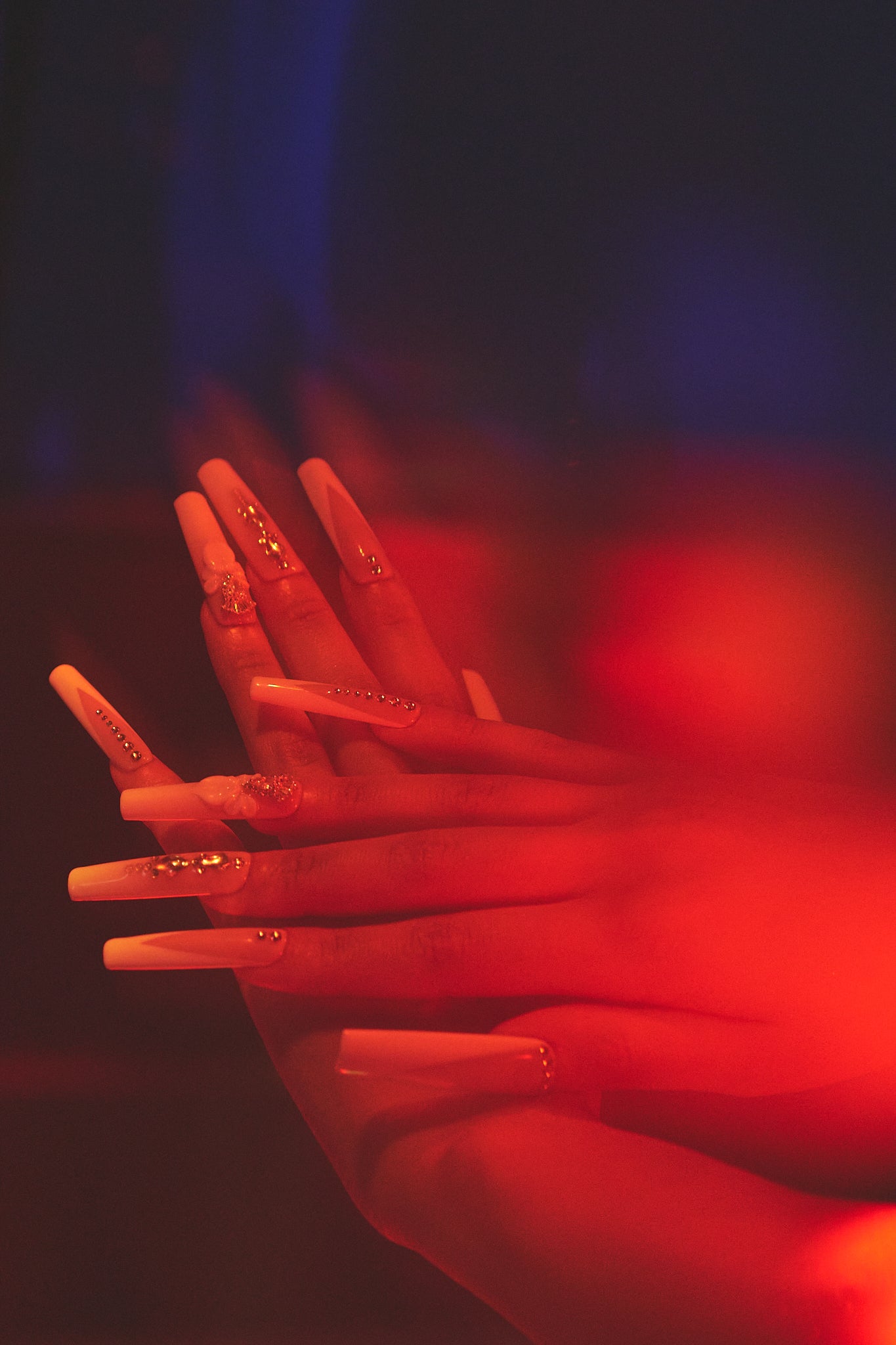 long square blush and white nails with gold accents glowing under red stage lighting with soft shadows