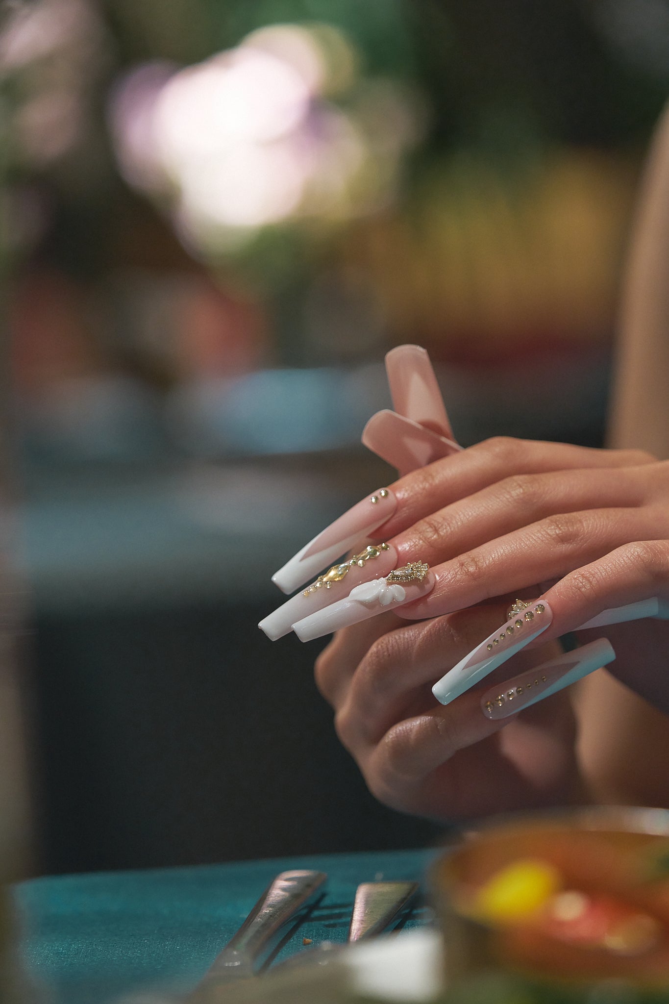 long square nails in blush pink and white with gold embellishments and 3D bow shown on hands over a blurred tablescape background