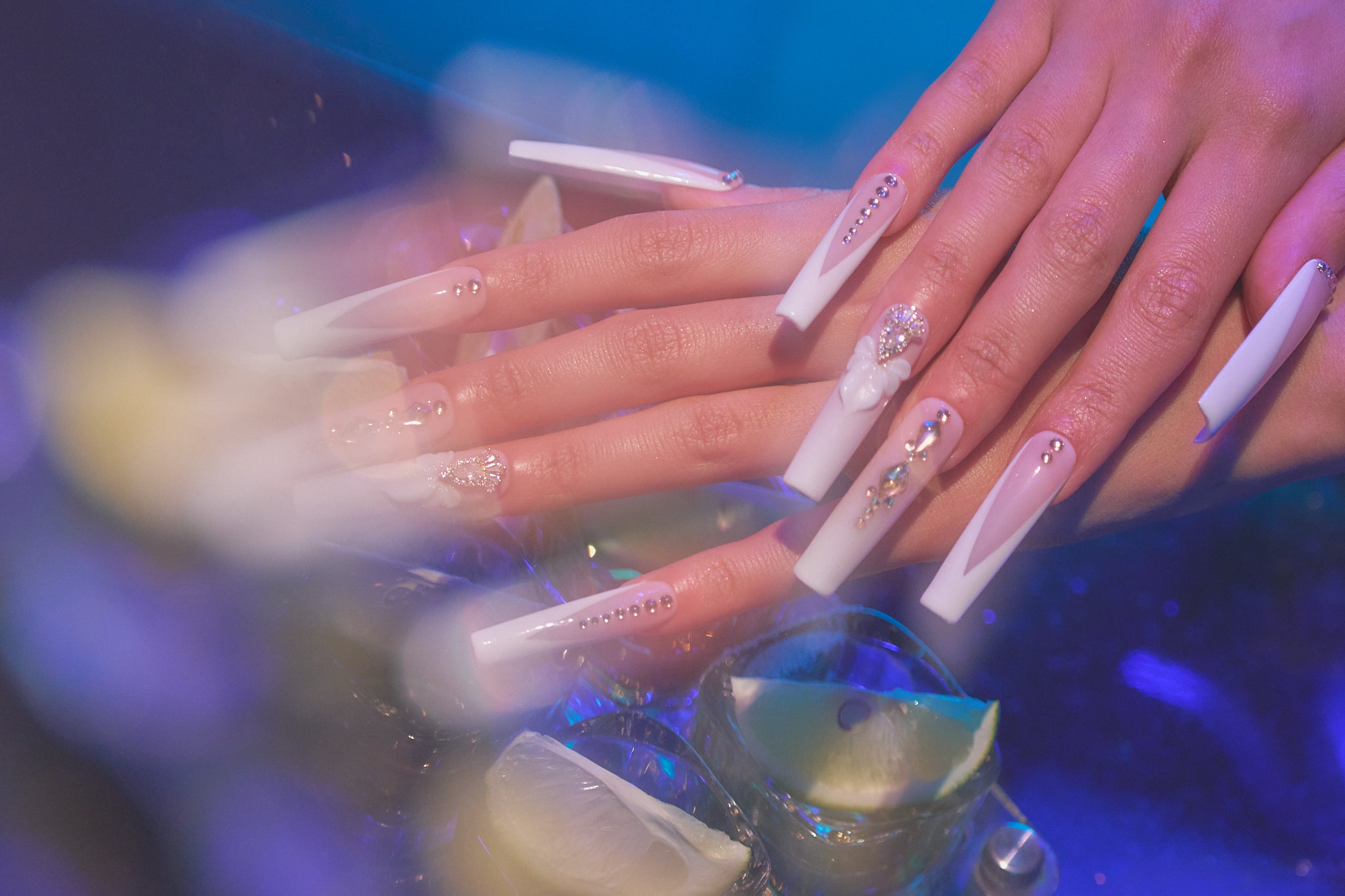 close-up of hands with long, decorated nails holding glass with blue liquid and lime slice, under soft purple-blue lighting