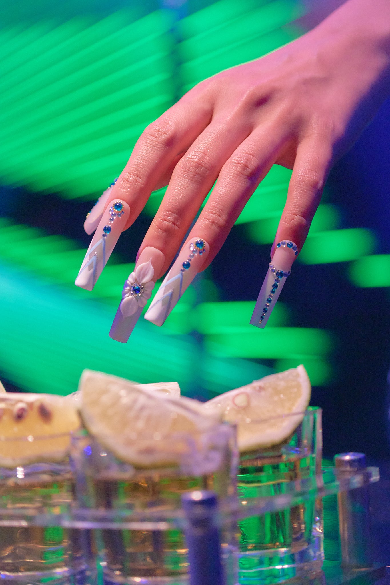 Vertical close-up of hand with ‘Frost Waltz’ long square press-on nails reaching toward tequila glasses with lime, under vibrant green lighting.