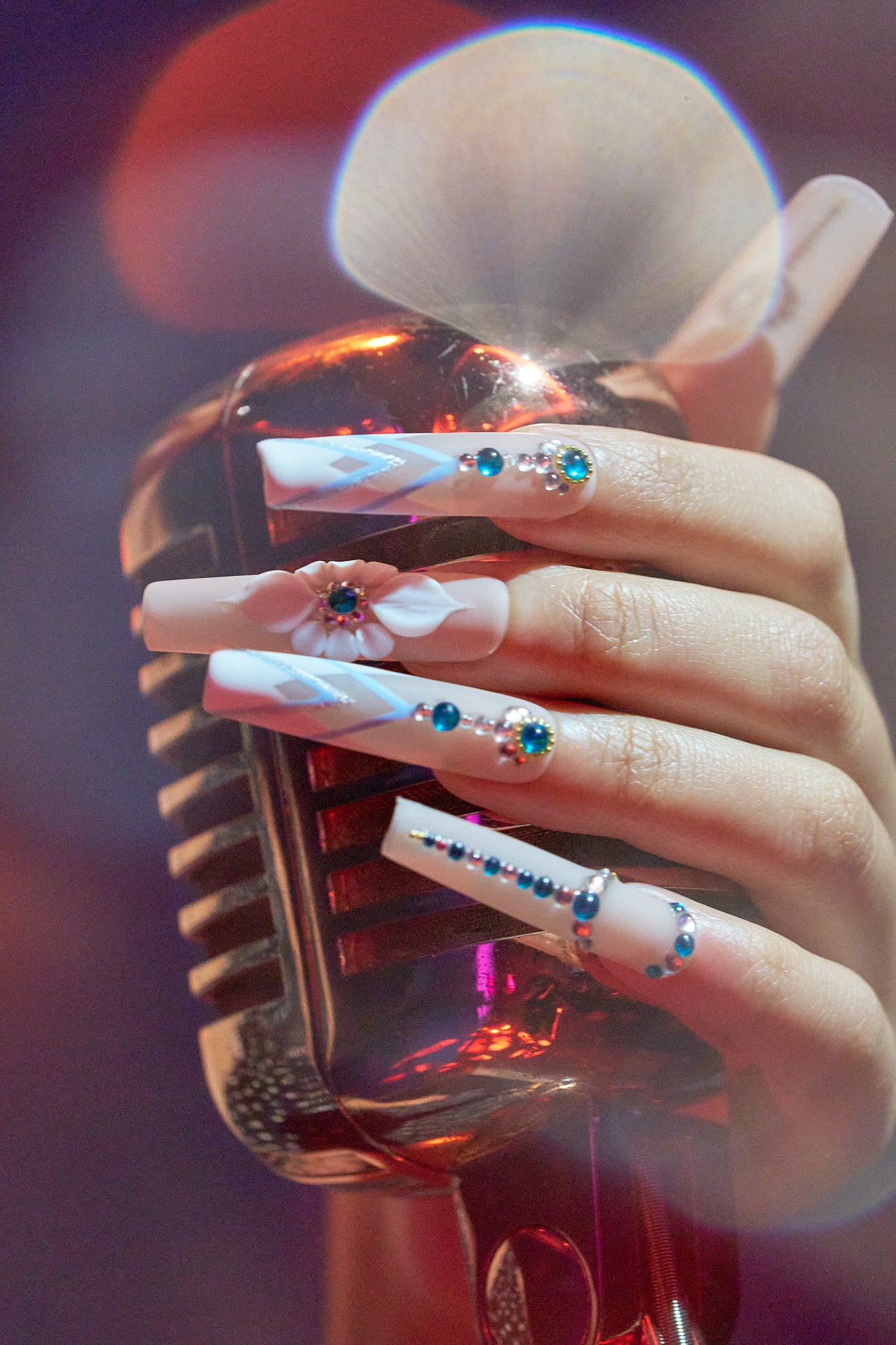 Hand wearing ‘Frost Waltz’ long square press-on nails with rhinestones and floral art, holding a vintage-style microphone under soft concert lights.