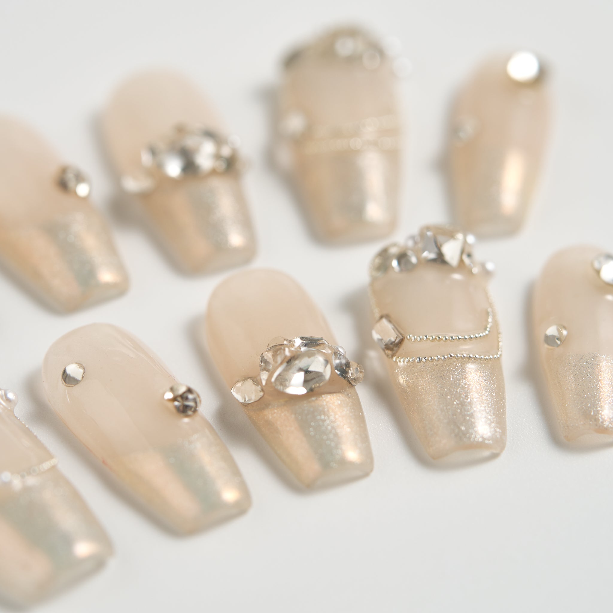Close-up of Gilded Heiress press-on nails in beige with rhinestones and delicate metallic chains, displayed on a white background.