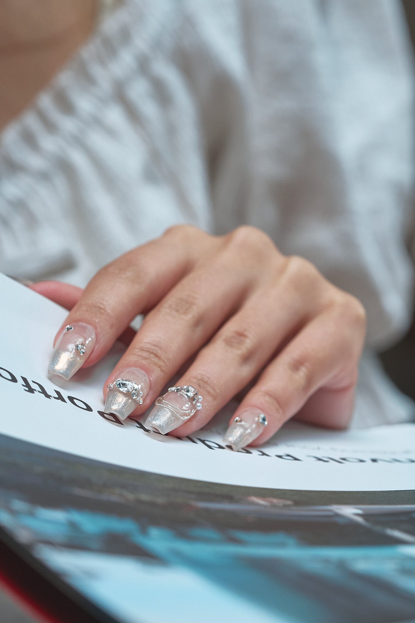 Hand with Gilded Heiress beige rhinestone press-on nails resting on an open magazine, with a soft blurred background.