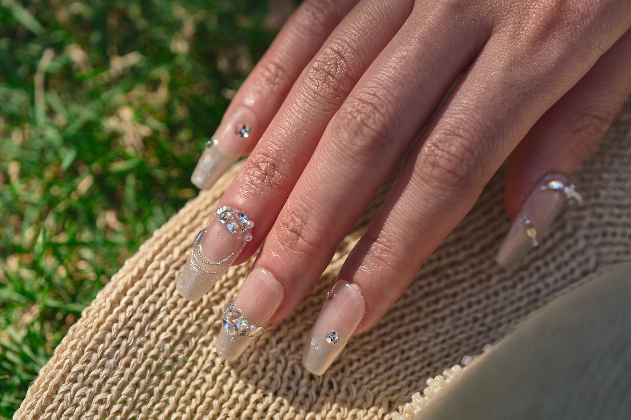 Close-up of a hand with Gilded Heiress decorative rhinestone nails resting on a beige knit fabric, with a blurred grass background.