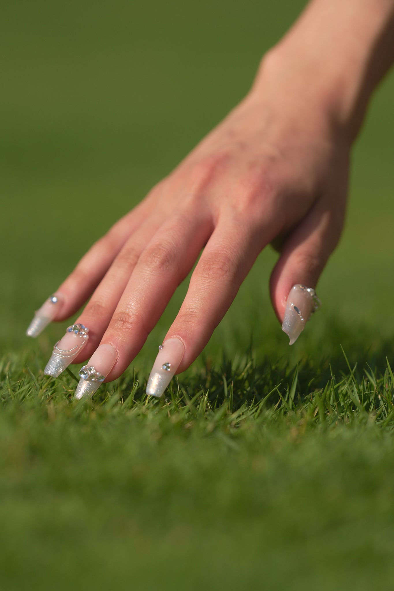 Close-up of a hand with Gilded Heiress decorative rhinestone nails touching grass, with a blurred green grass background.