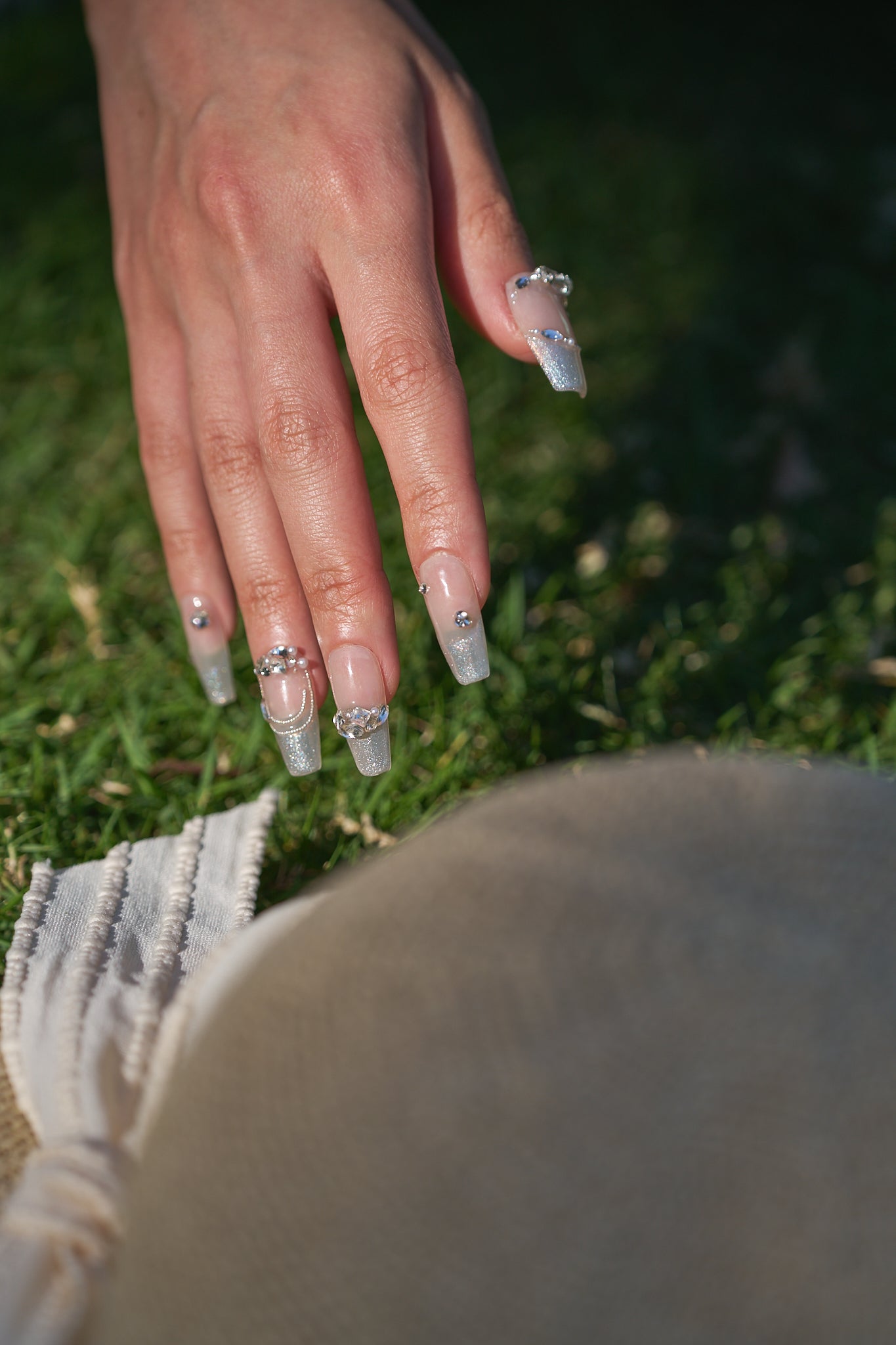 Close-up of a hand with Gilded Heiress decorative rhinestone nails resting on a grassy outdoor background, with a beige hat partially visible.