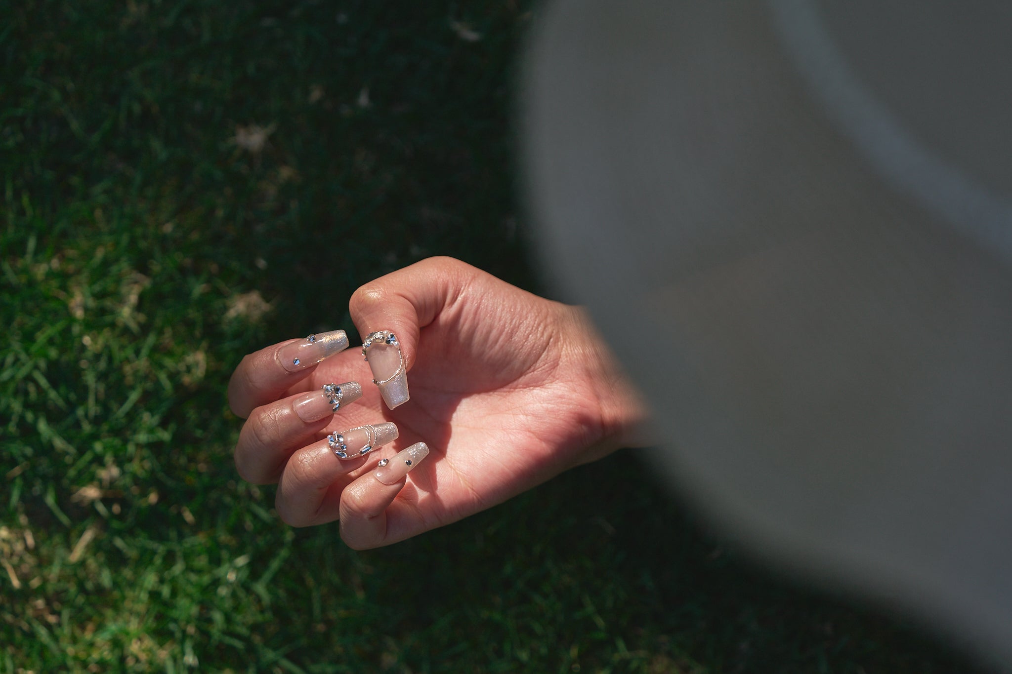 Close-up of a hand with Gilded Heiress decorative rhinestone nails outdoors on grass.