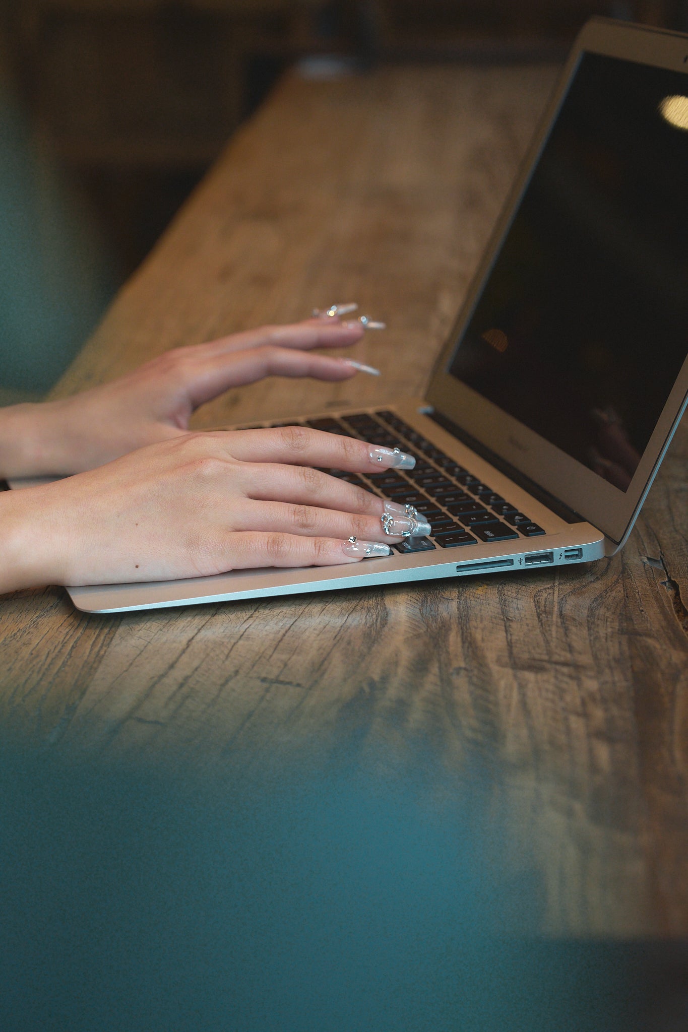 Hands with Gilded Heiress beige rhinestone press-on nails typing on a laptop at a wooden table.