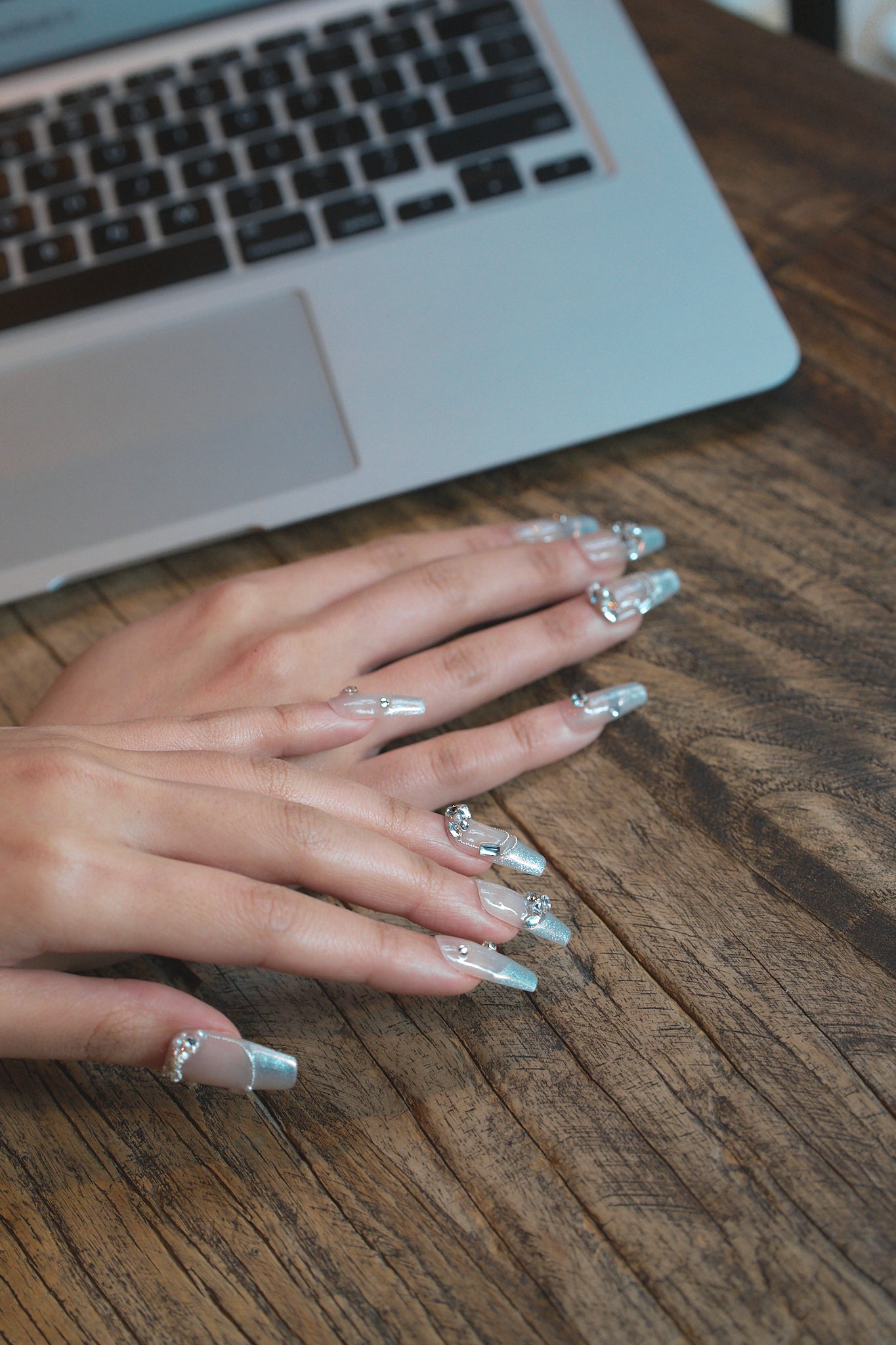 Close-up of hands with Gilded Heiress rhinestone nails resting on a wooden desk next to a laptop.