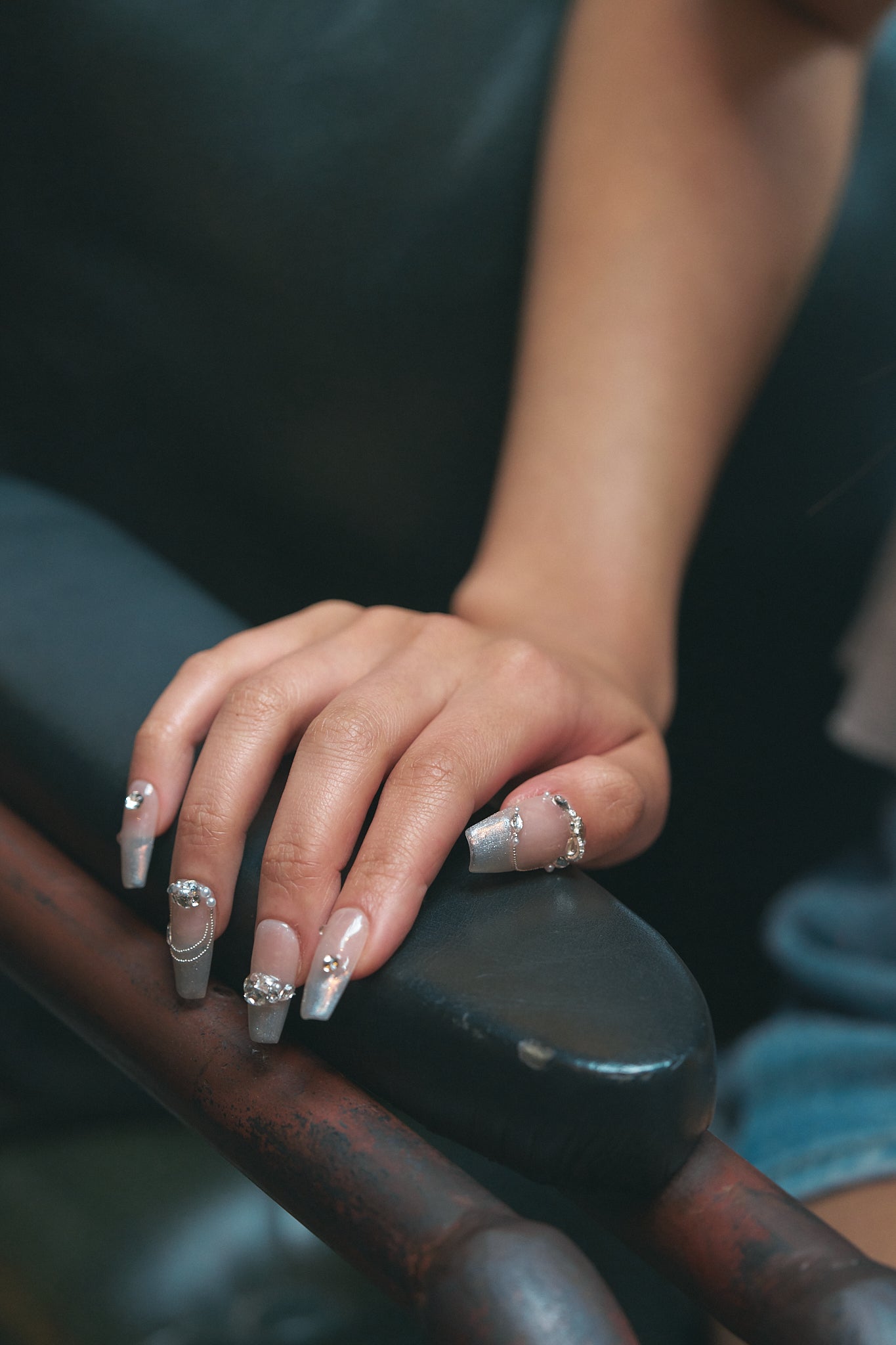 Hand wearing Gilded Heiress beige rhinestone press-on nails and rings, resting on a dark chair.