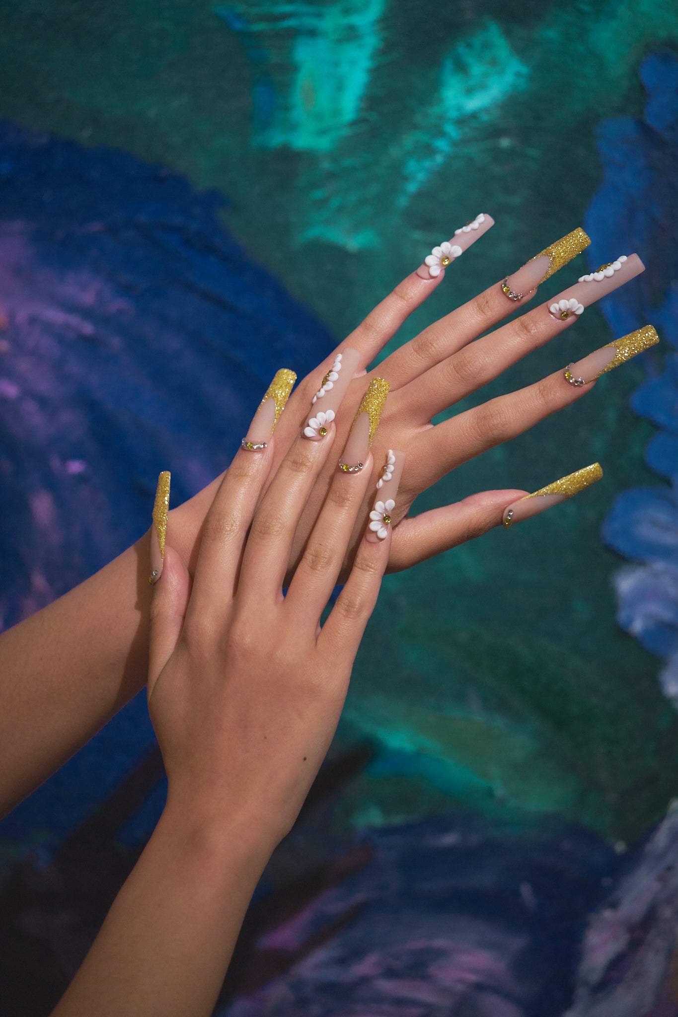 Hands wearing ‘Golden Bloom’ long square press-on nails with gold glitter and 3D daisies, posed in front of an abstract blue-green painted background.