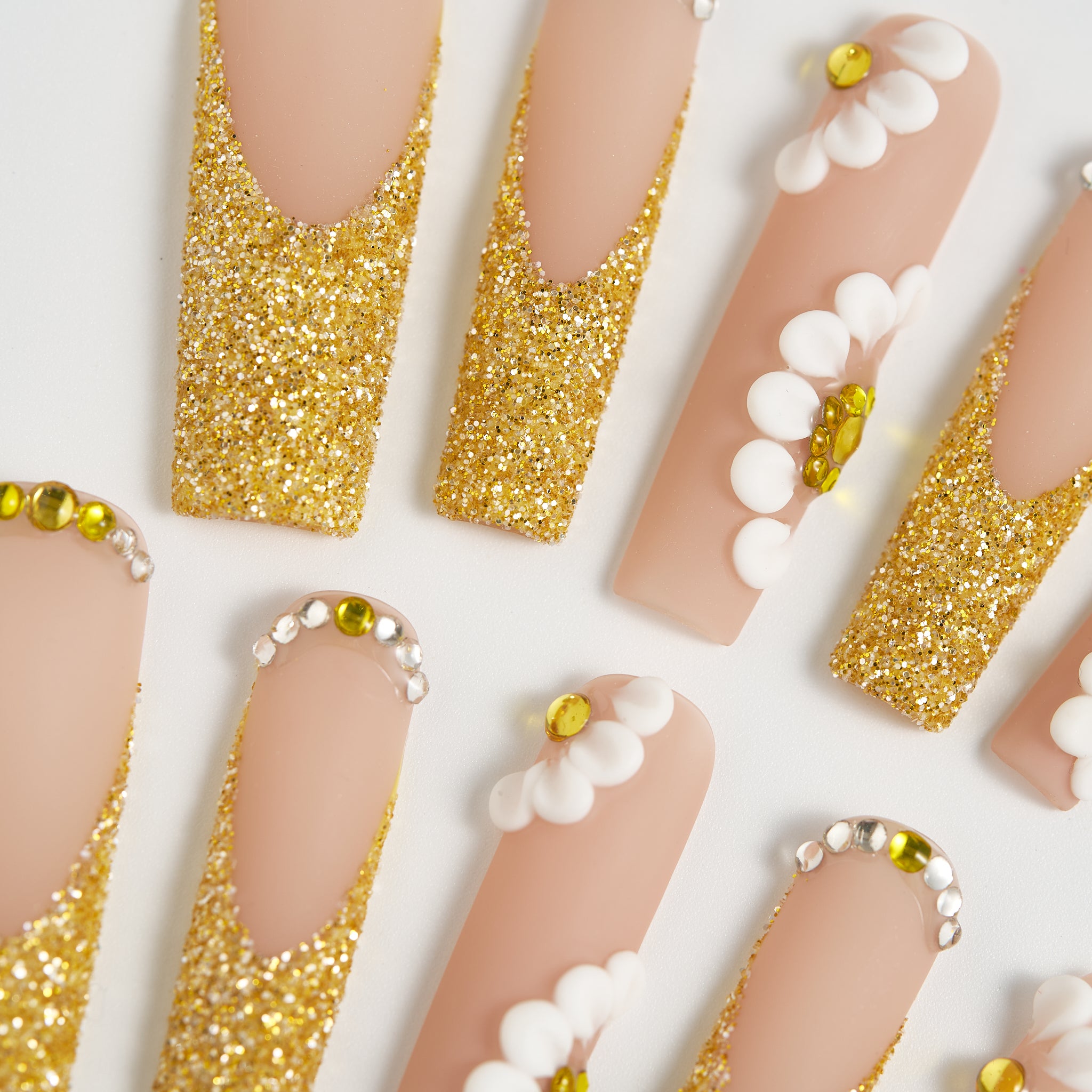 Close-up of ‘Golden Bloom’ long square press-on nails with gold glitter tips, white 3D daisies, and gem accents on a beige base.