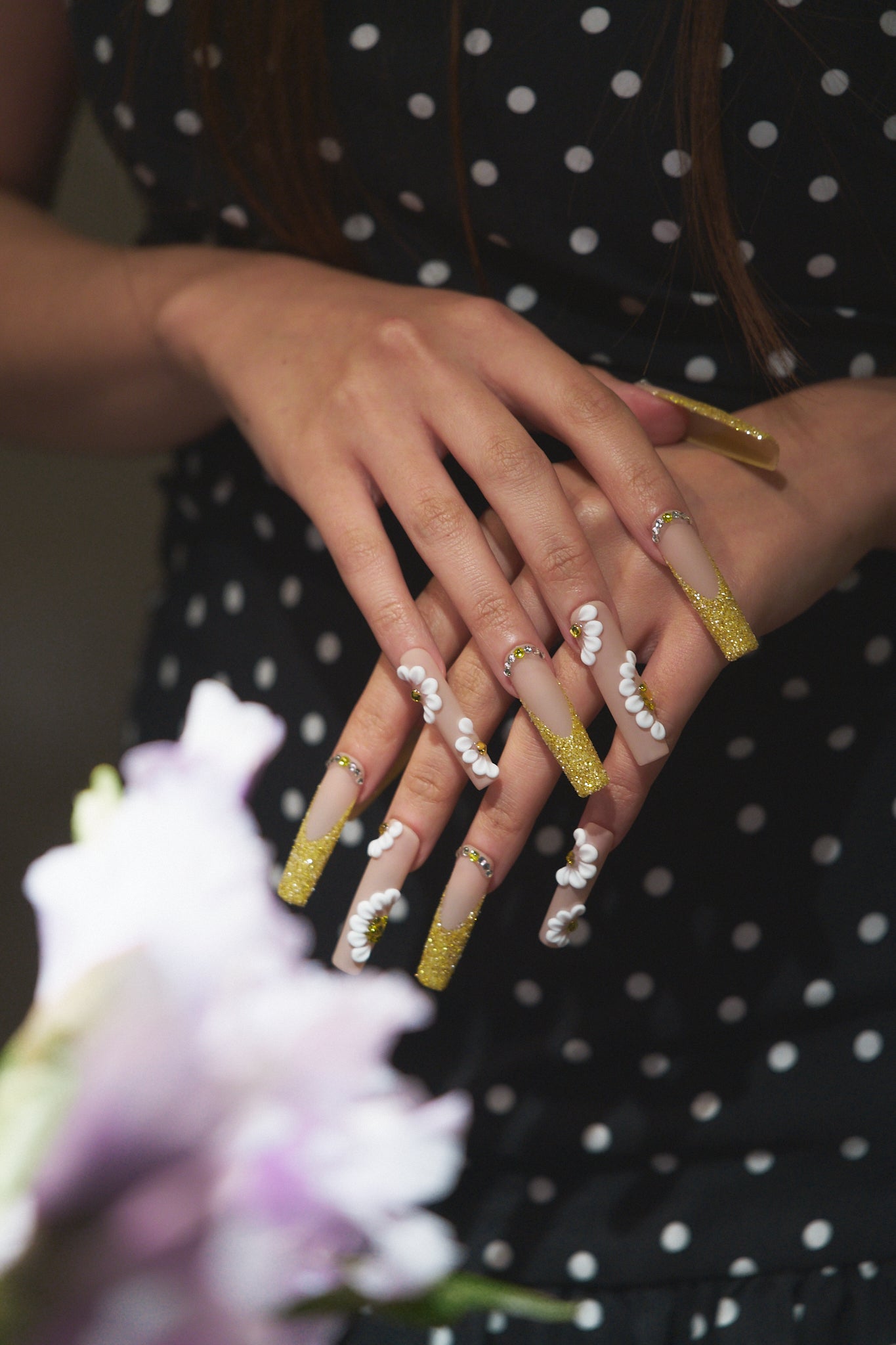Hands wearing ‘Golden Bloom’ long square press-on nails with gold glitter and white daisy accents, posed over a bouquet while wearing a black polka dot dress.
