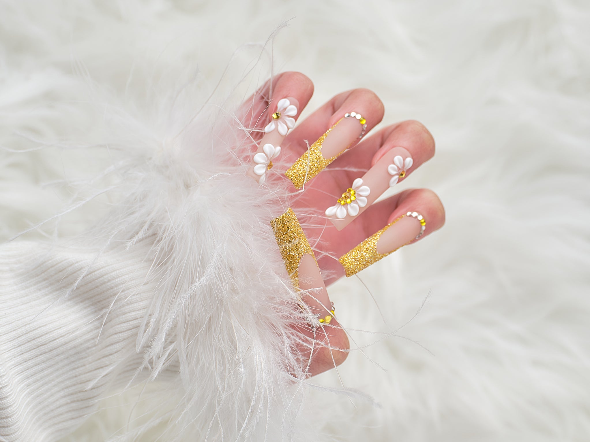 Full hand view of ‘Golden Bloom’ long square press-on nails with gold glitter and 3D daisy accents, styled with white knit and faux fur.