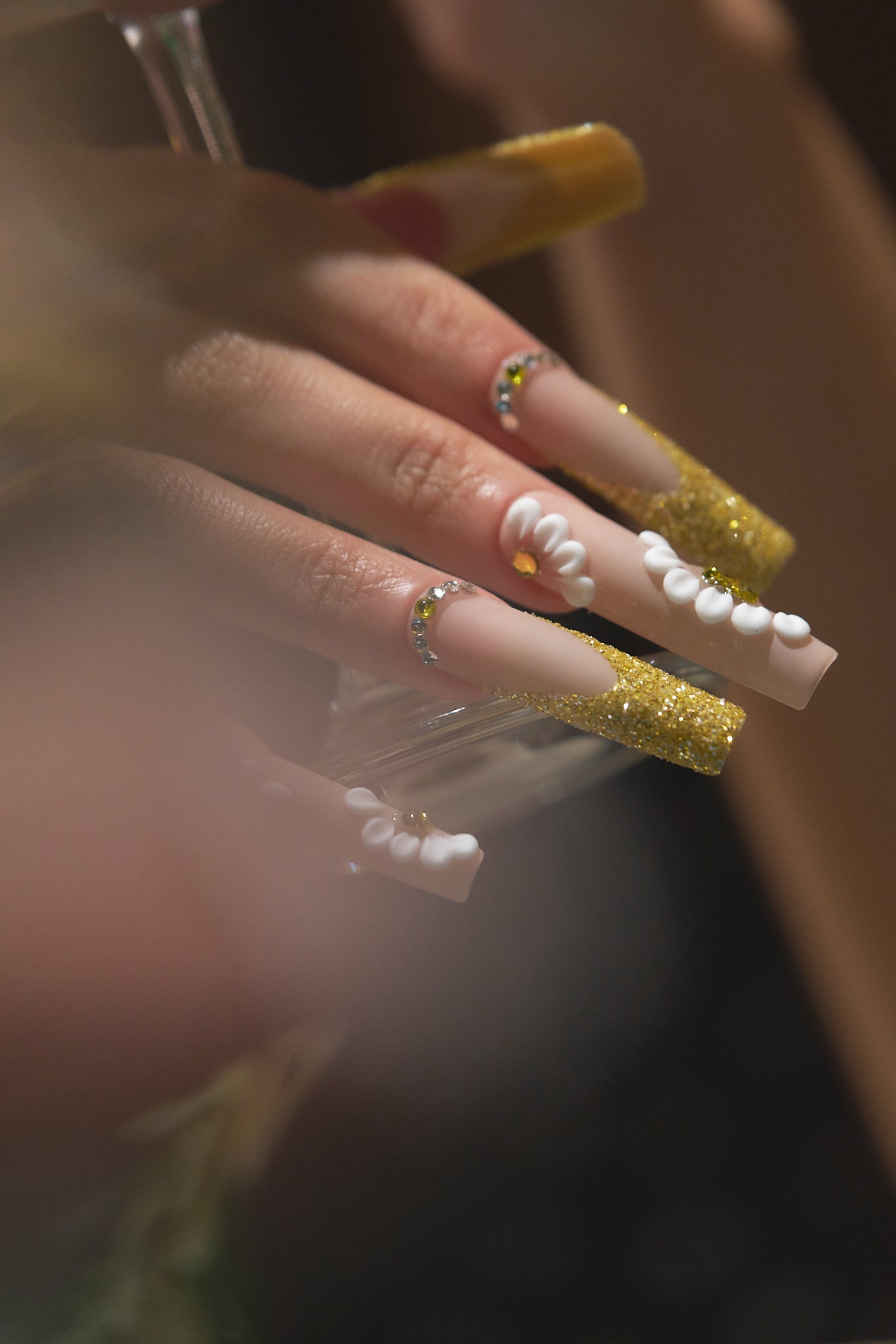 Close-up of hand with ‘Golden Bloom’ long square press-on nails holding a glass, featuring gold glitter tips and white daisy designs under soft lighting.