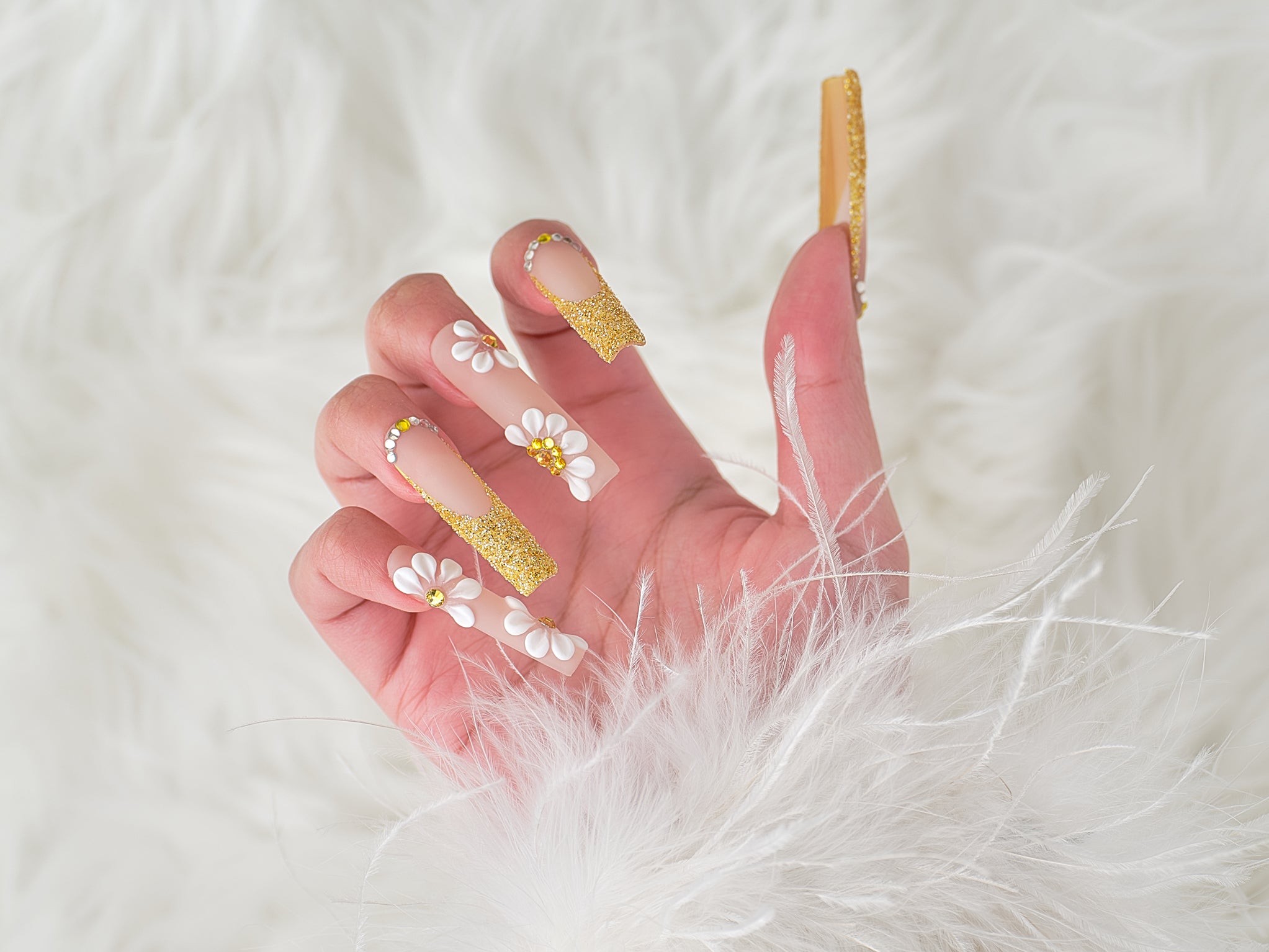 Side view of hand wearing ‘Golden Bloom’ long square press-on nails with gold glitter tips and 3D white daisy designs, posed against a fluffy white background.