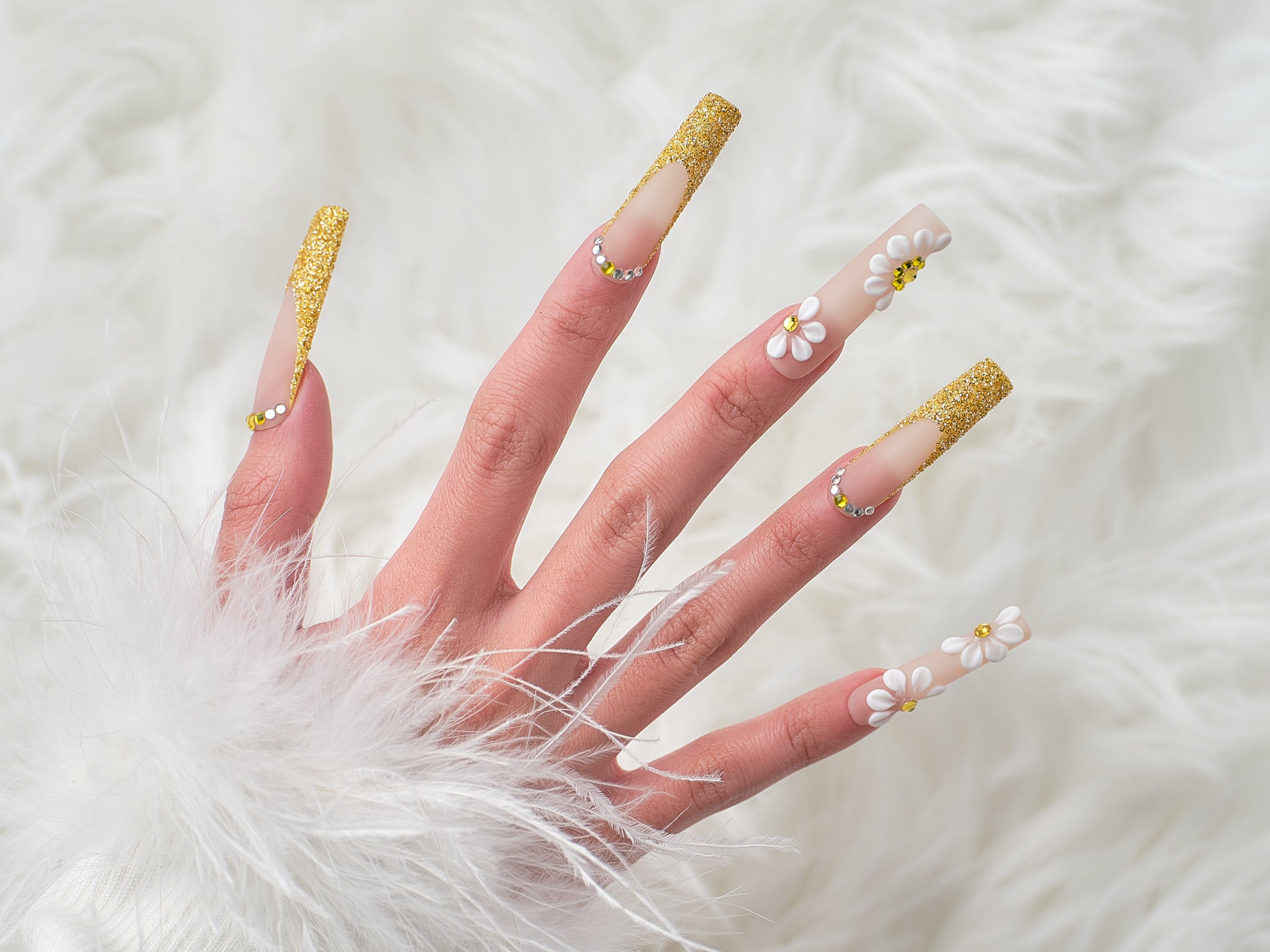 Hand wearing ‘Golden Bloom’ long square press-on nails with gold glitter tips and 3D white daisy accents, posed over a white faux fur background.
