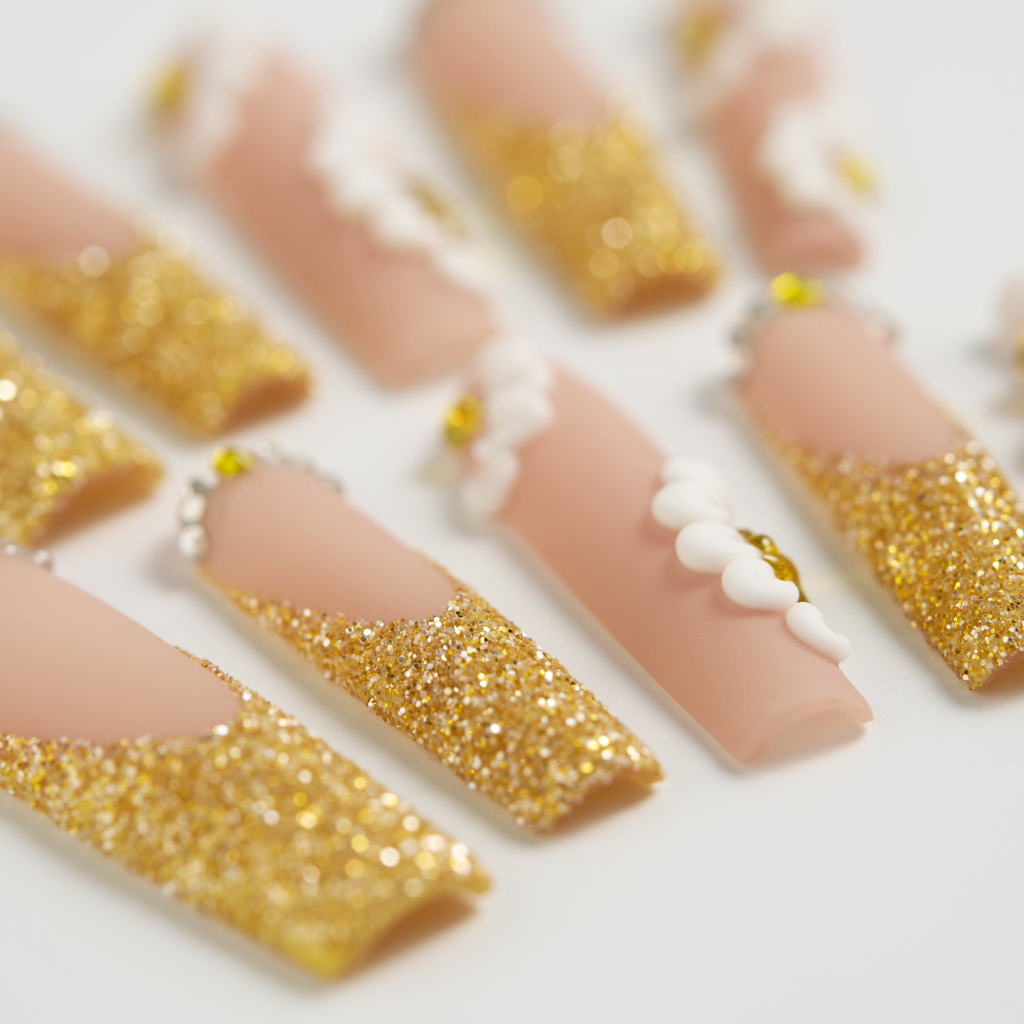 Depth-focused macro of ‘Golden Bloom’ long square press-on nails featuring gold glitter, white 3D daisies, and gemstone detailing on a beige base.