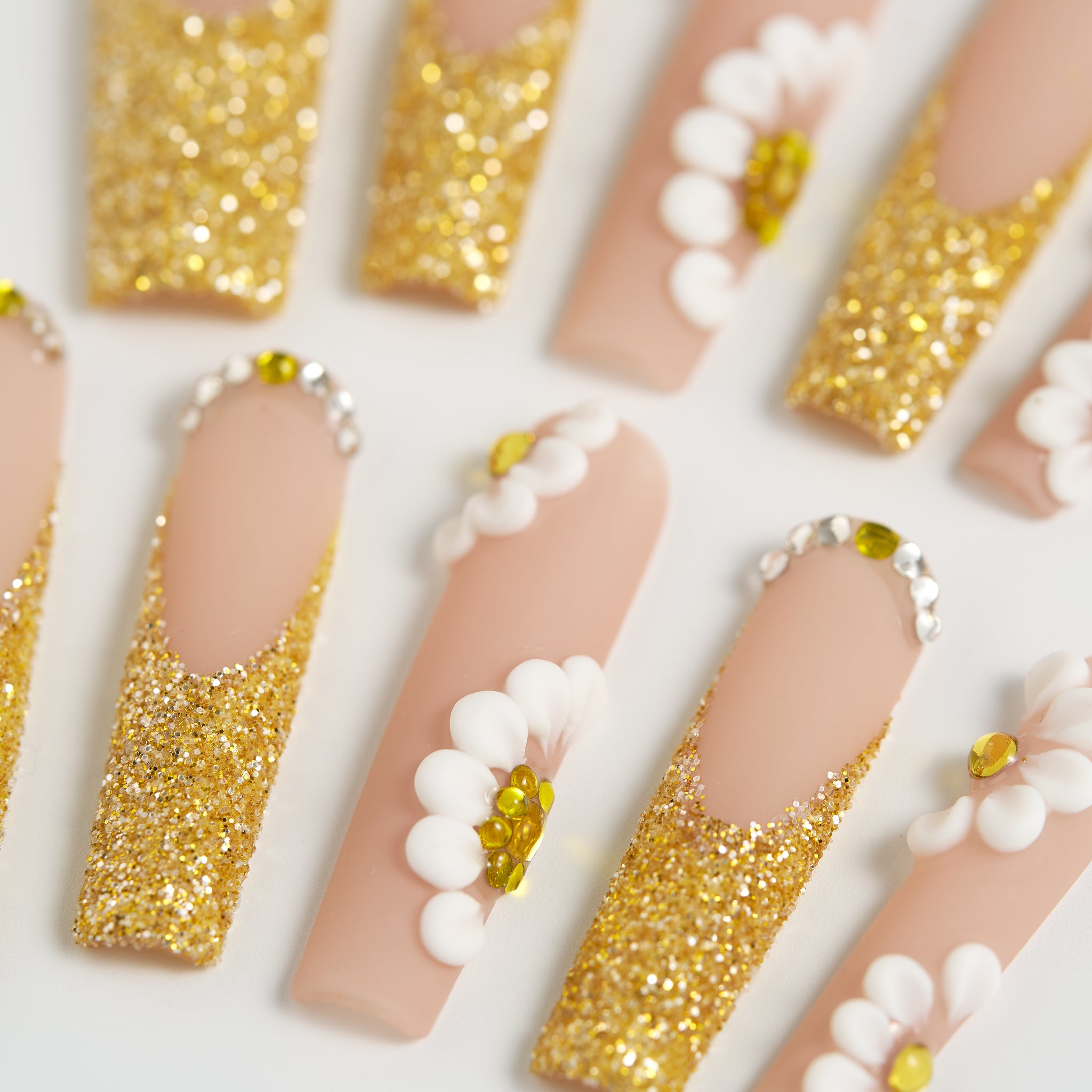 Balanced macro shot of ‘Golden Bloom’ long square press-on nails with gold glitter, 3D white daisies, and gemstone accents on a beige base.