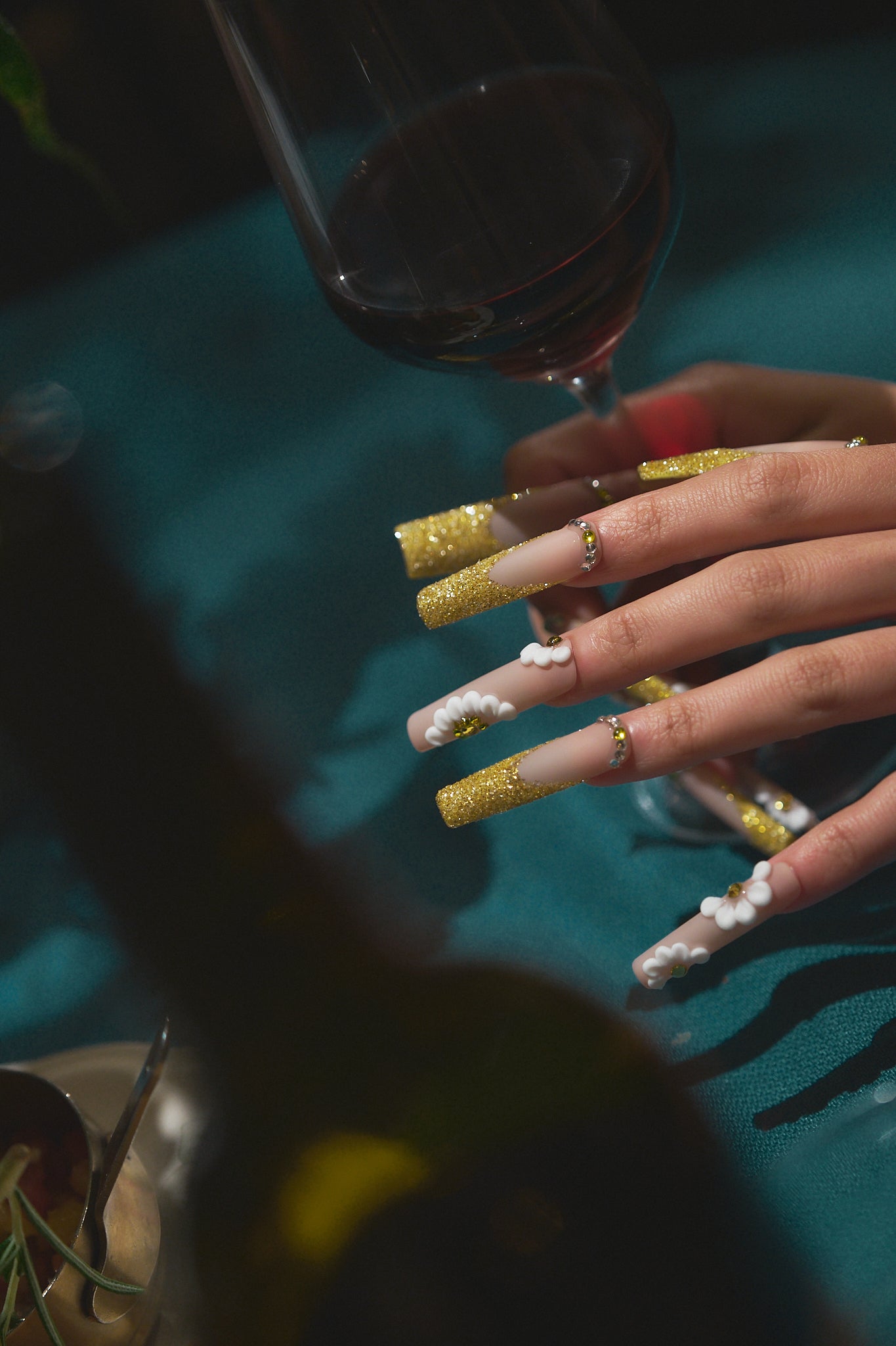 long square nails in gold glitter and 3D white detail holding a red wine glass on dark background