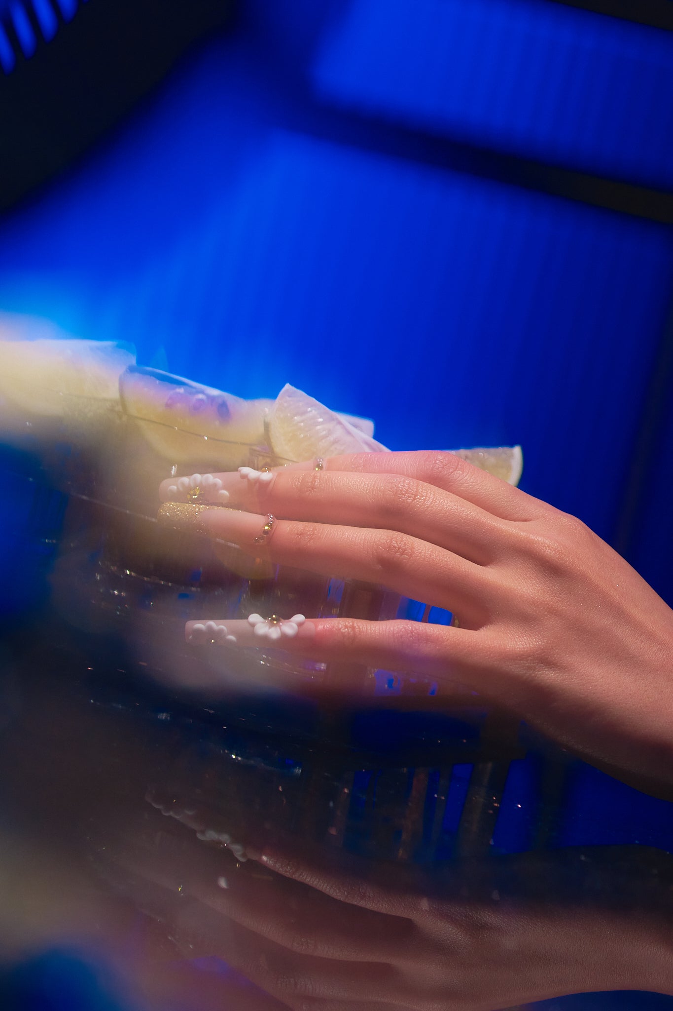 long square nails in gold glitter with 3D white florals holding a glass with lemon slices under blue lighting