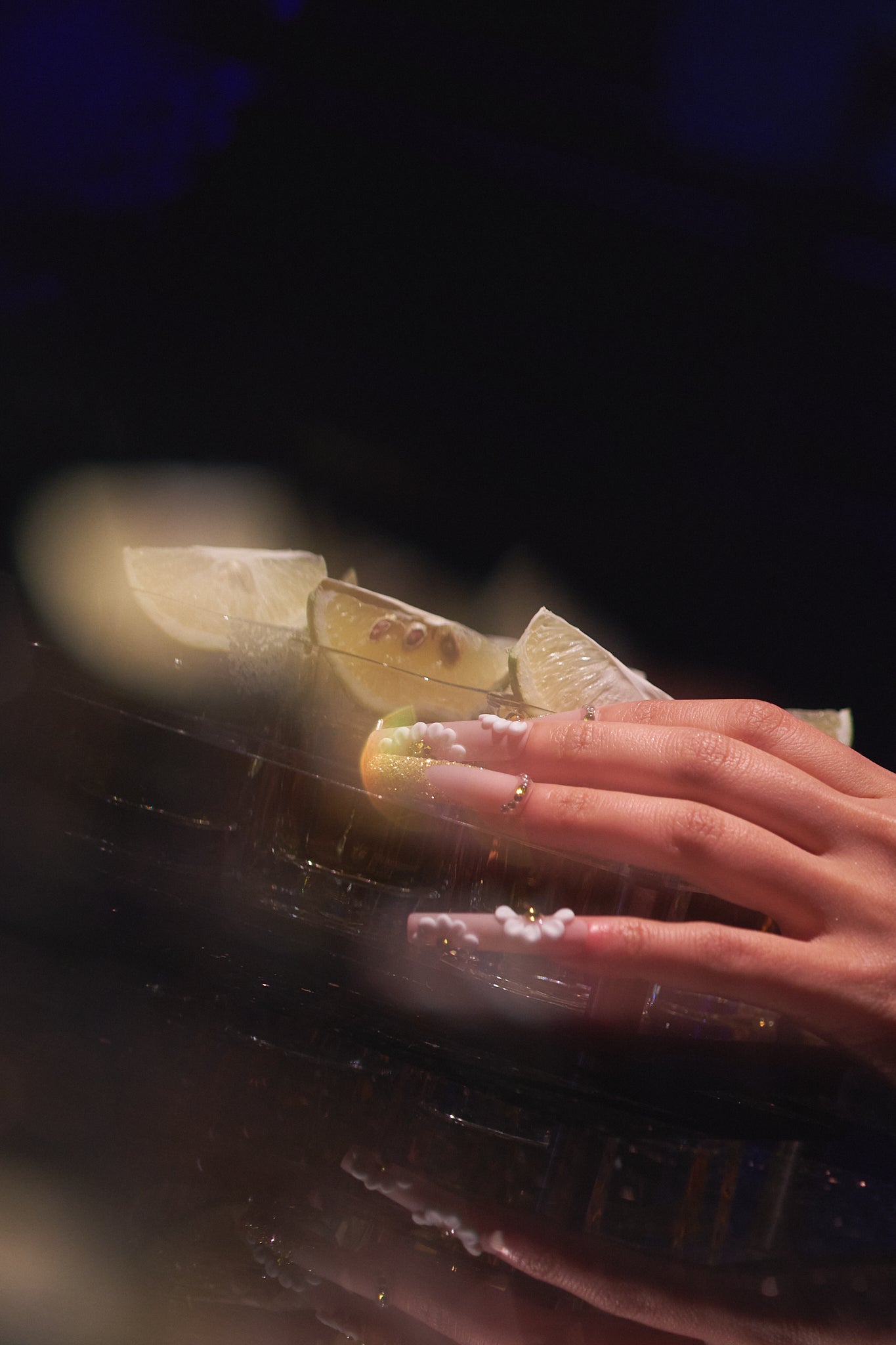 long square nails in gold glitter with 3D white floral details holding a glass with lemon slices on a dark background