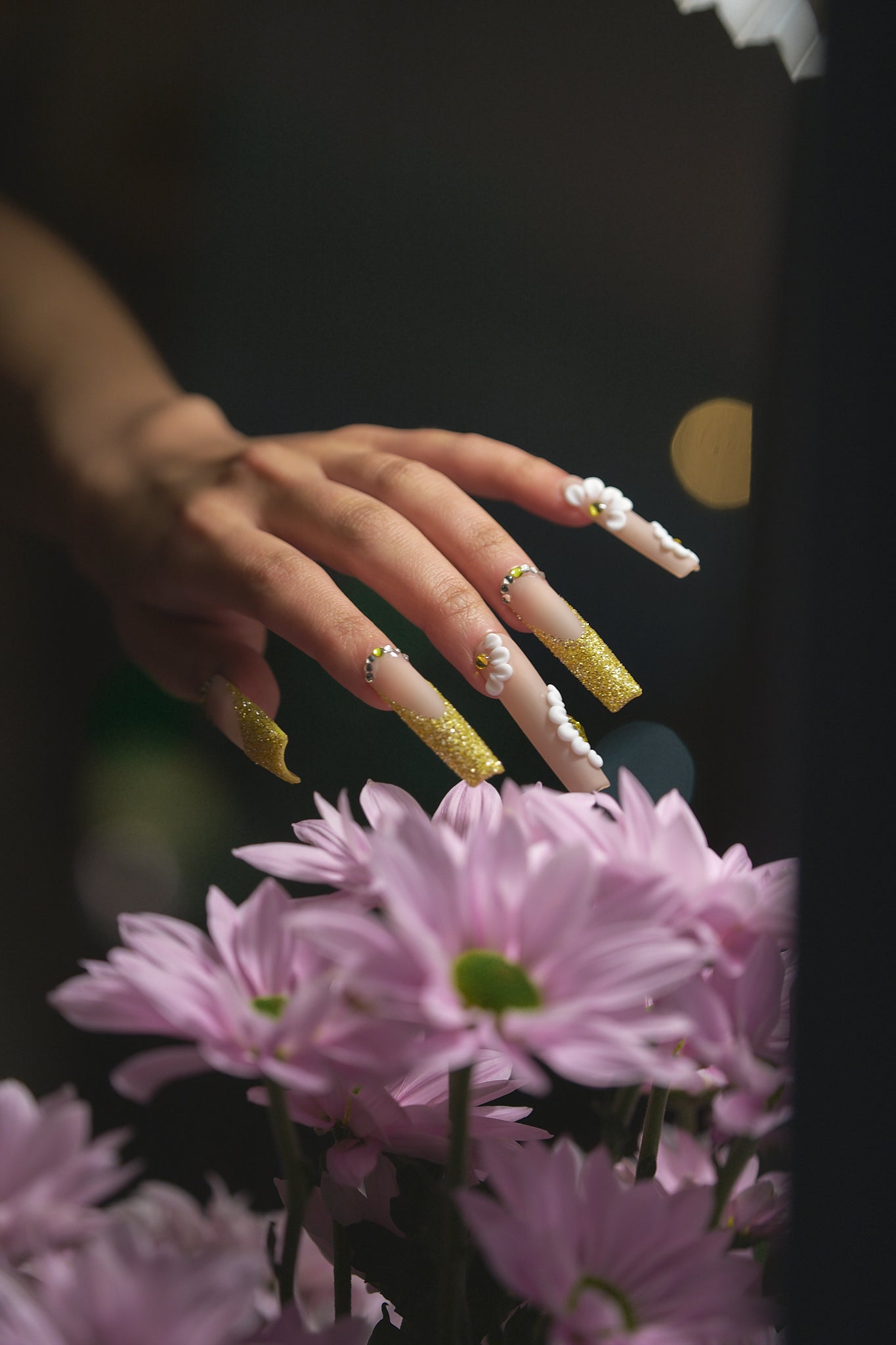 long square nails in gold glitter with 3D white florals reaching toward pink flowers on dark background