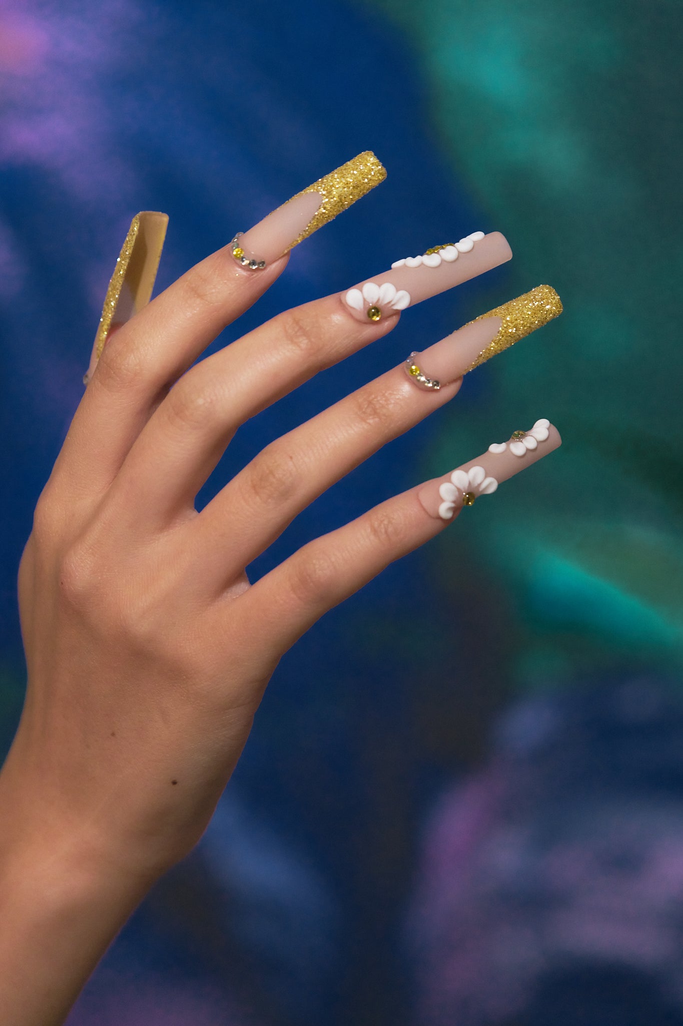 long square nails in gold glitter with 3D white floral accents shown against a colorful blurred backdrop