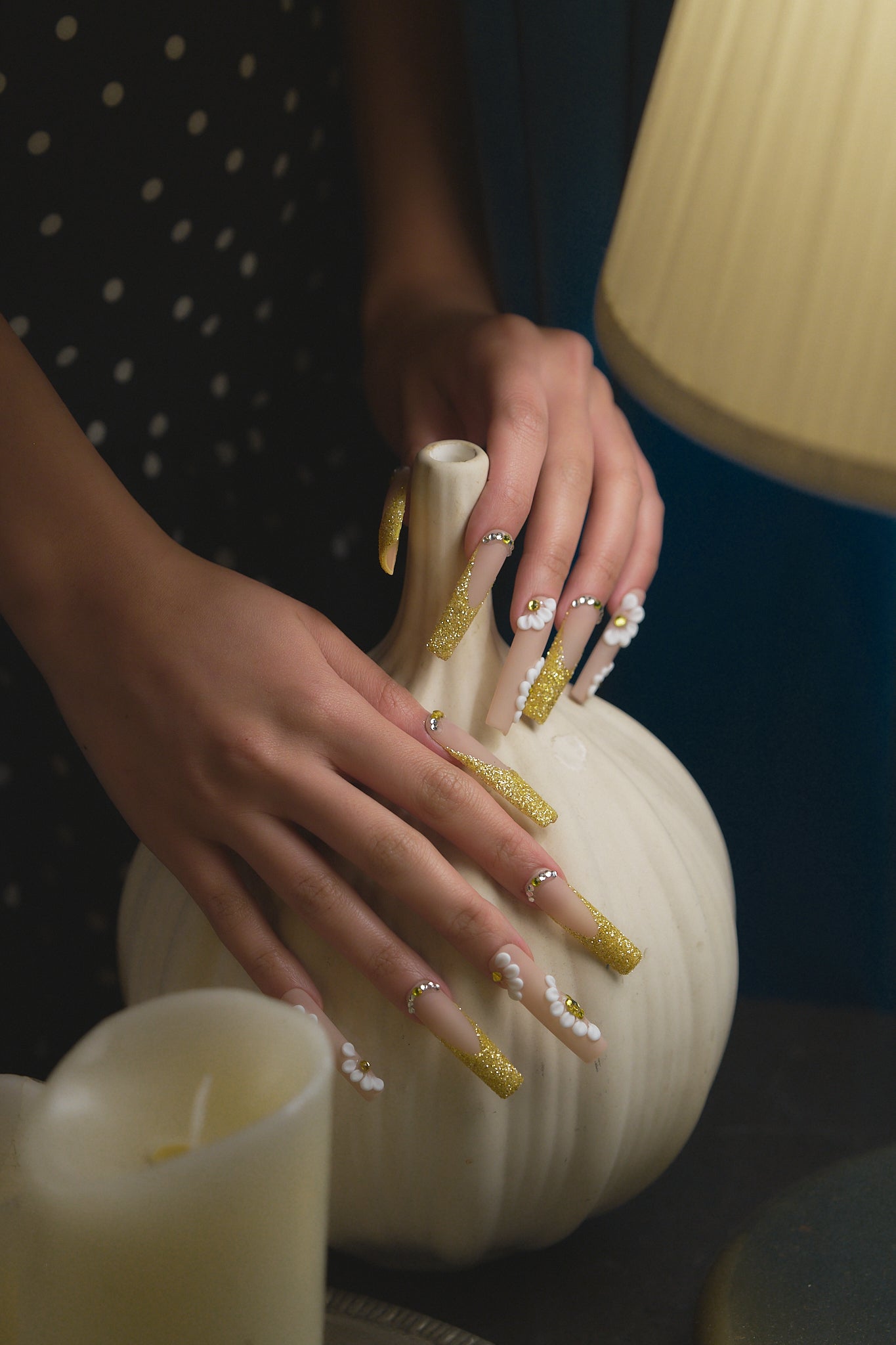 long square nails in gold glitter and white 3D flowers touching a white pumpkin on dark background