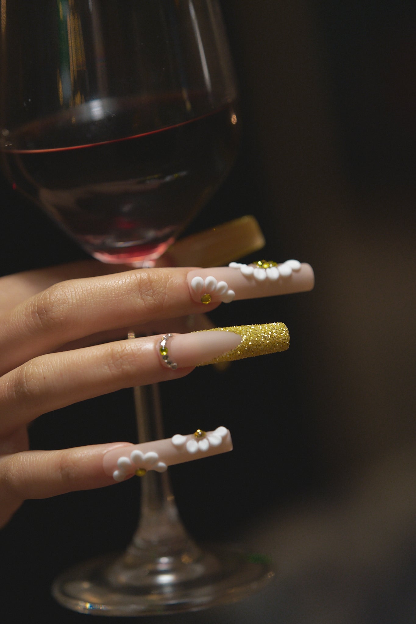 close-up of long square nails in gold glitter with 3D white florals holding a red wine glass on dark background