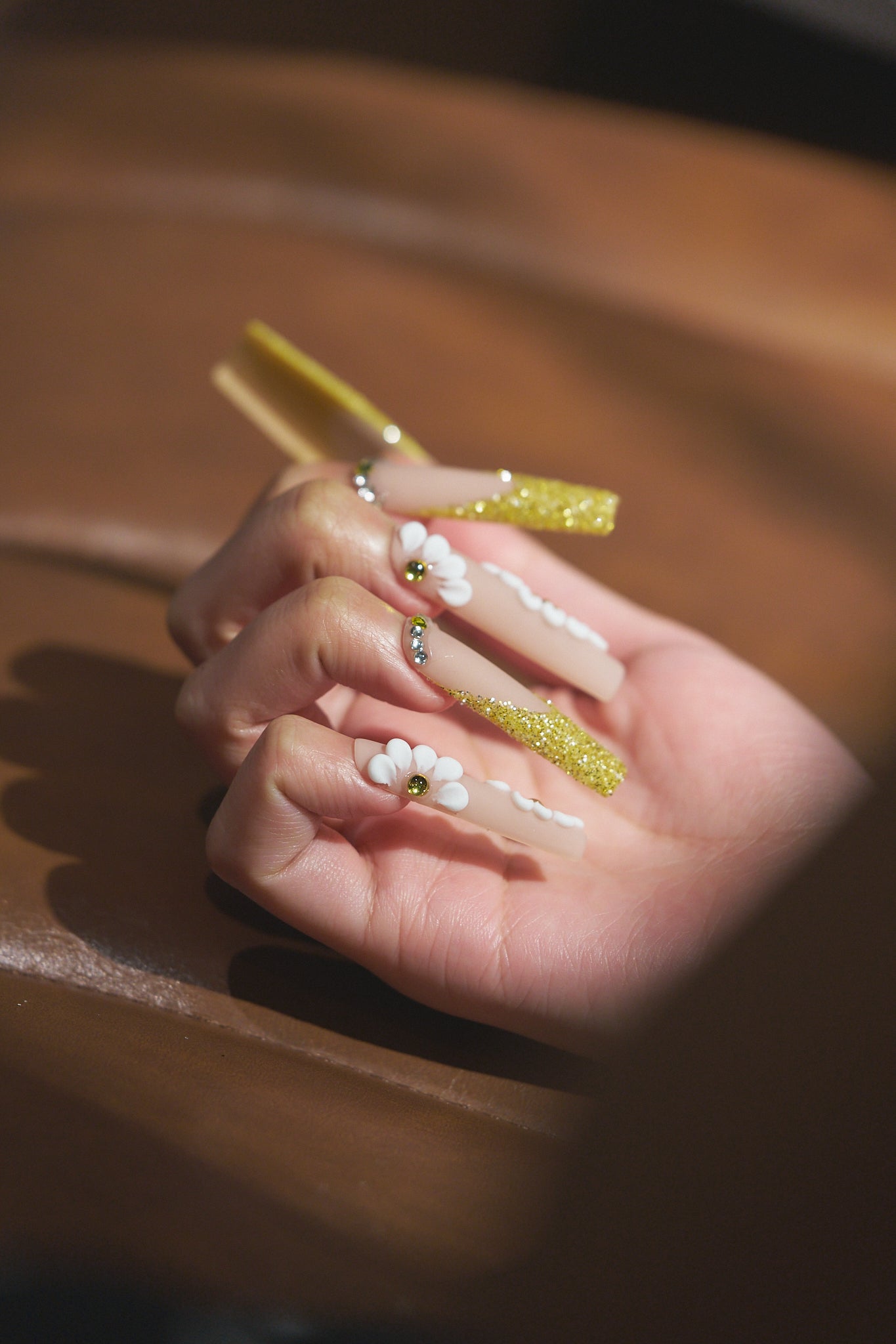 long square nails in gold glitter with 3D white florals resting on a wooden surface in warm light