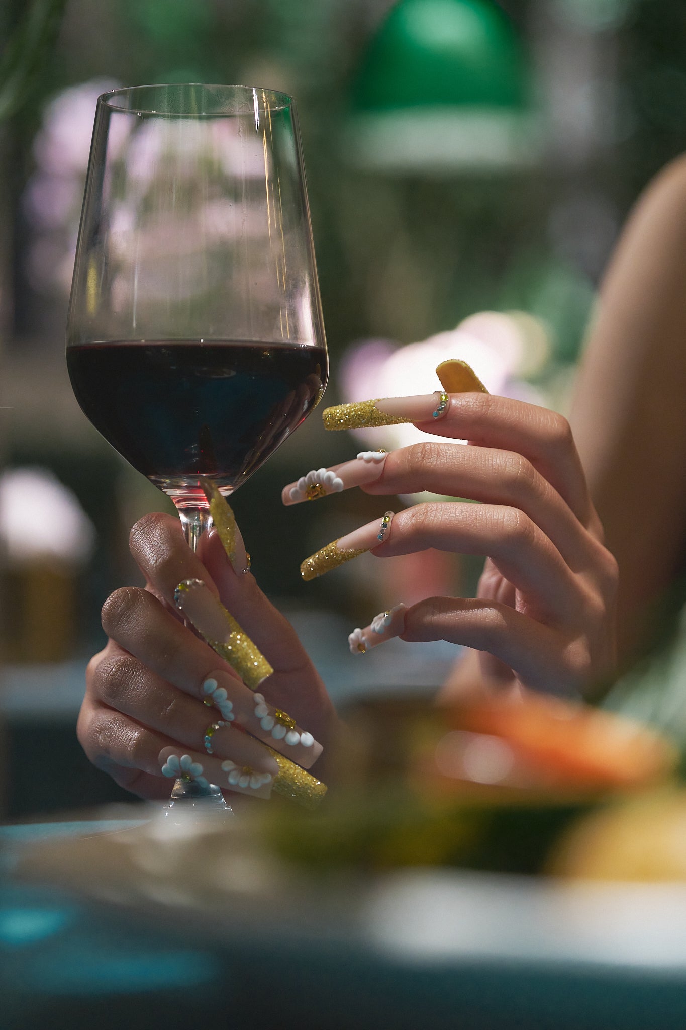 Hands wearing ‘Golden Bloom’ long square press-on nails holding a wine glass, with gold glitter and 3D white flowers in a soft daylight brunch setting.