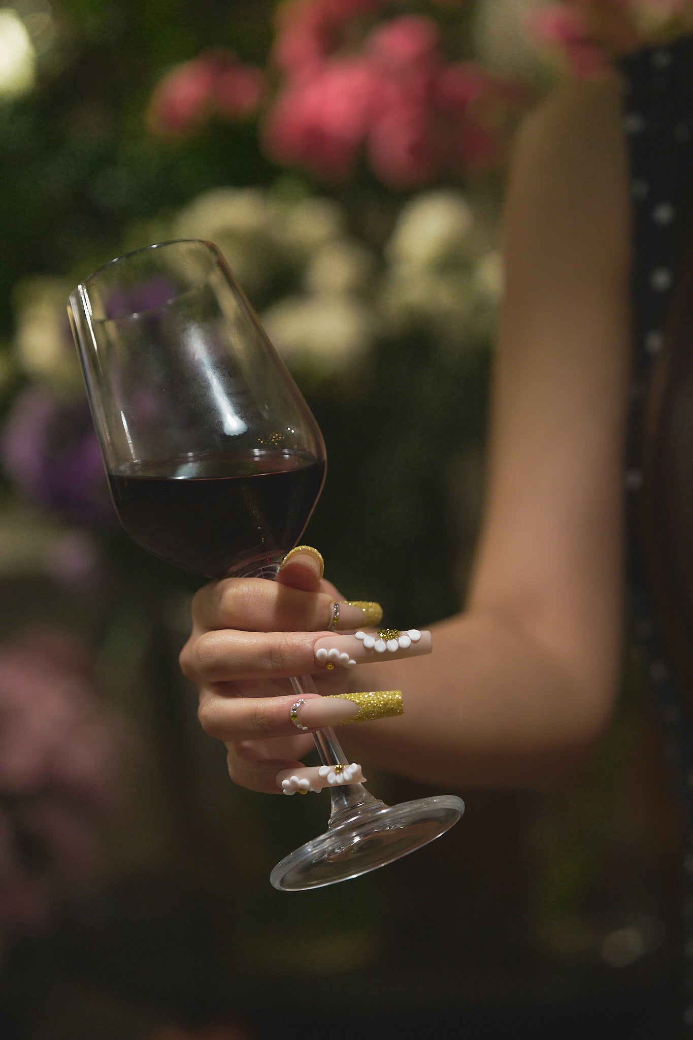 Hand with ‘Golden Bloom’ long square press-on nails holding a wine glass, featuring white floral nail art and gold glitter, set against a blurred floral background.