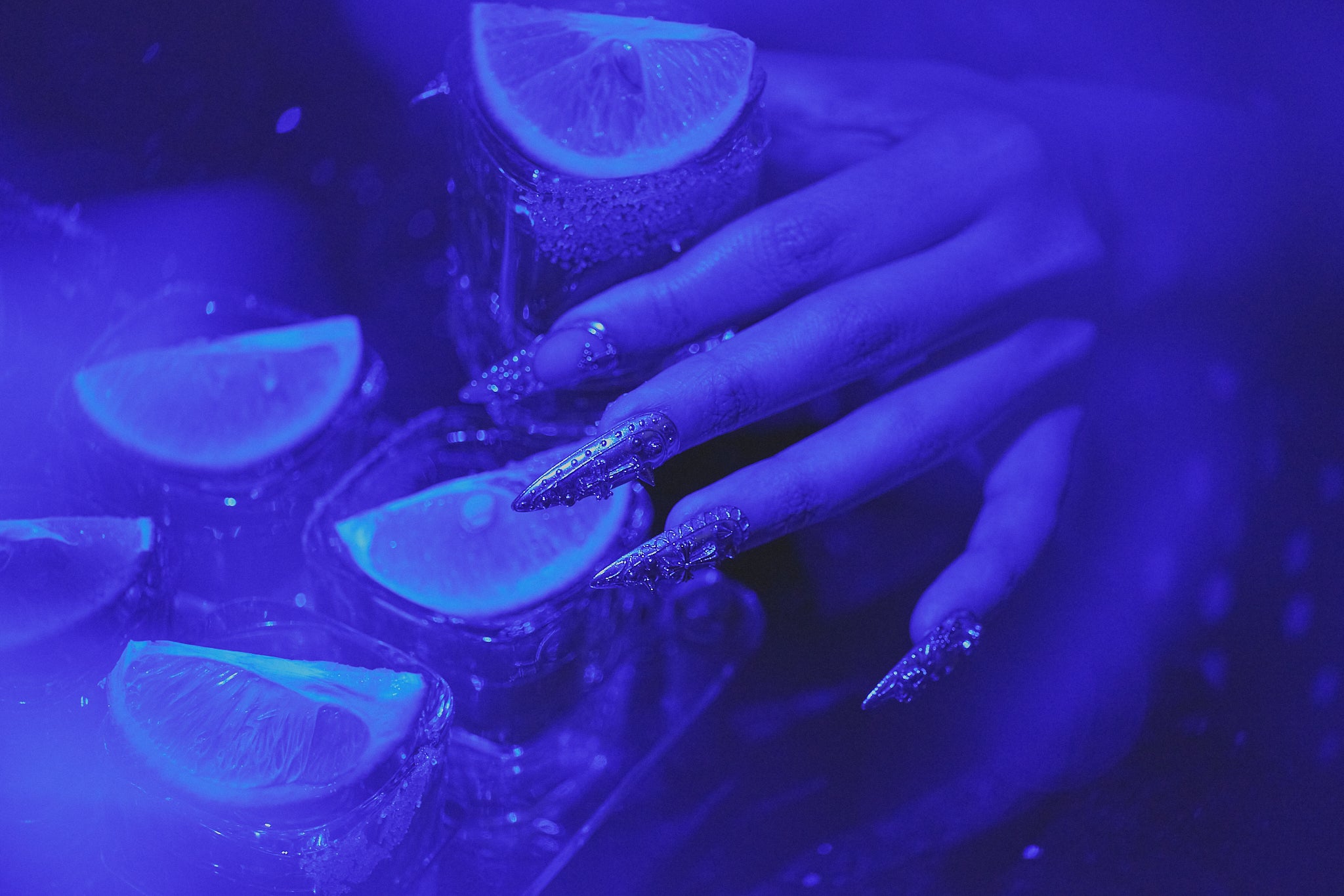 Hand wearing MiuNail “Chrome Reign” long glitter press-on nails, reaching toward glasses with lime slices under dramatic blue lighting.