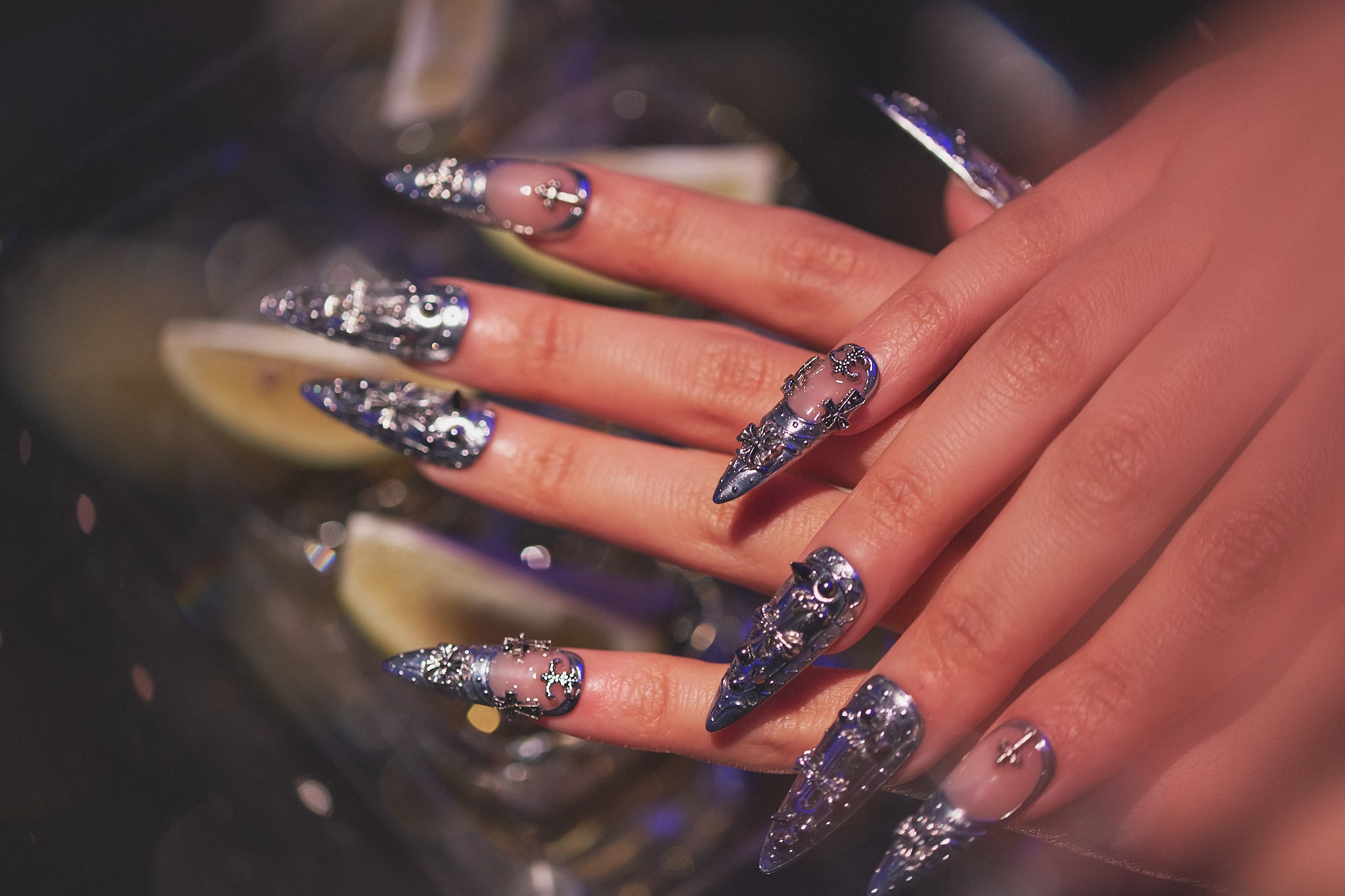 Hands with MiuNail Chrome Reign metallic press-on nails, dramatic silver gothic details, resting on a bar counter surrounded by tequila shots and lime, modern nightlife vibe.
