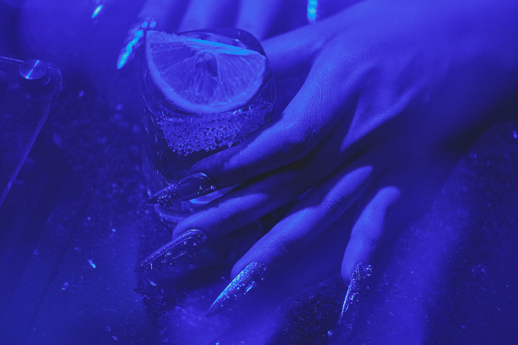 MiuNail Chrome Reign, the medium stiletto press-on nails in metallic chrome silver, hand holding tequila shot with lime under neon blue light, nightlife manicure inspiration.