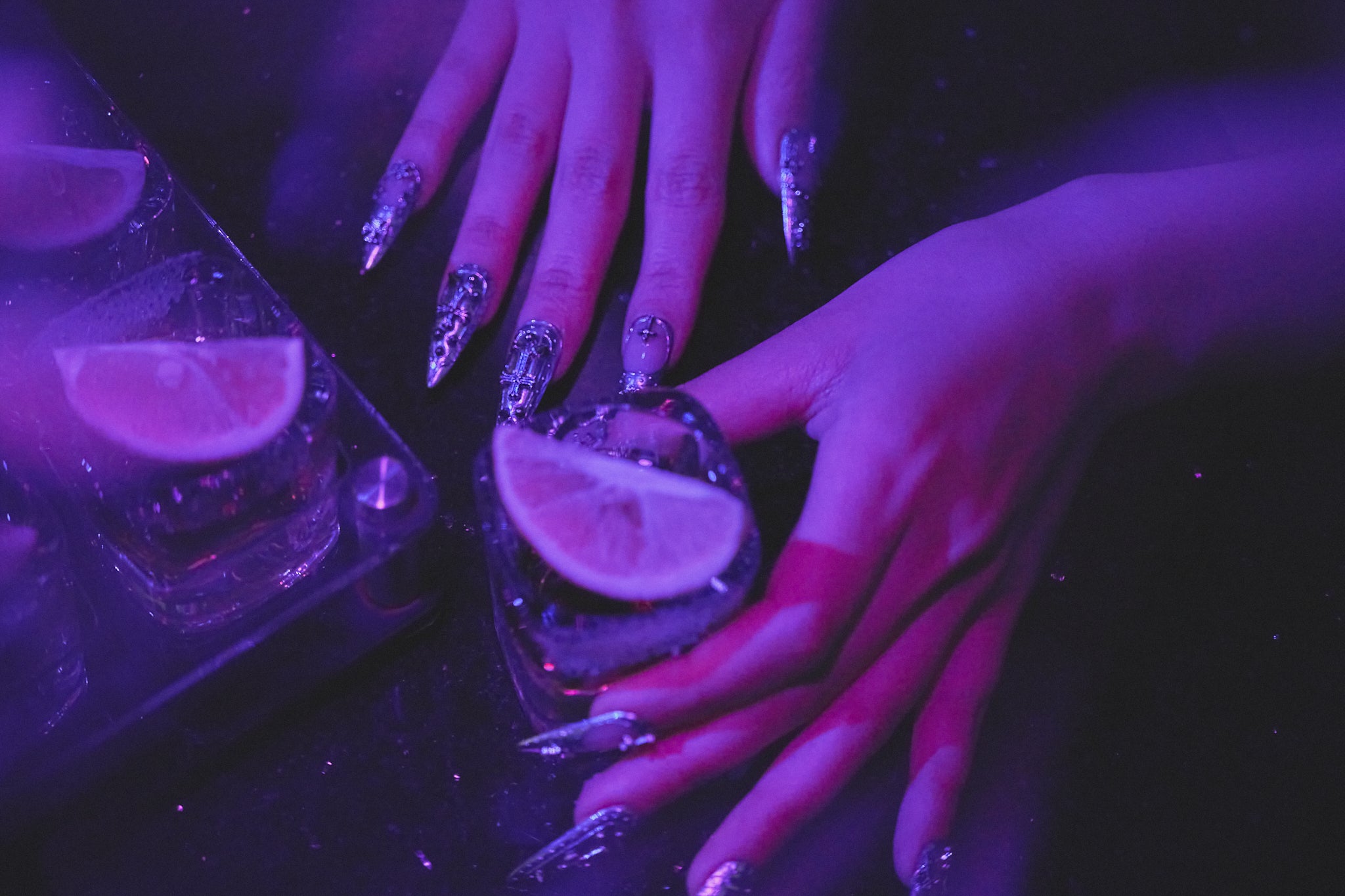 Chrome Reign, MiuNail press-on nails in metallic silver, both hands with stiletto manicure holding tequila shots with lime, vibrant purple nightlife bar scene.