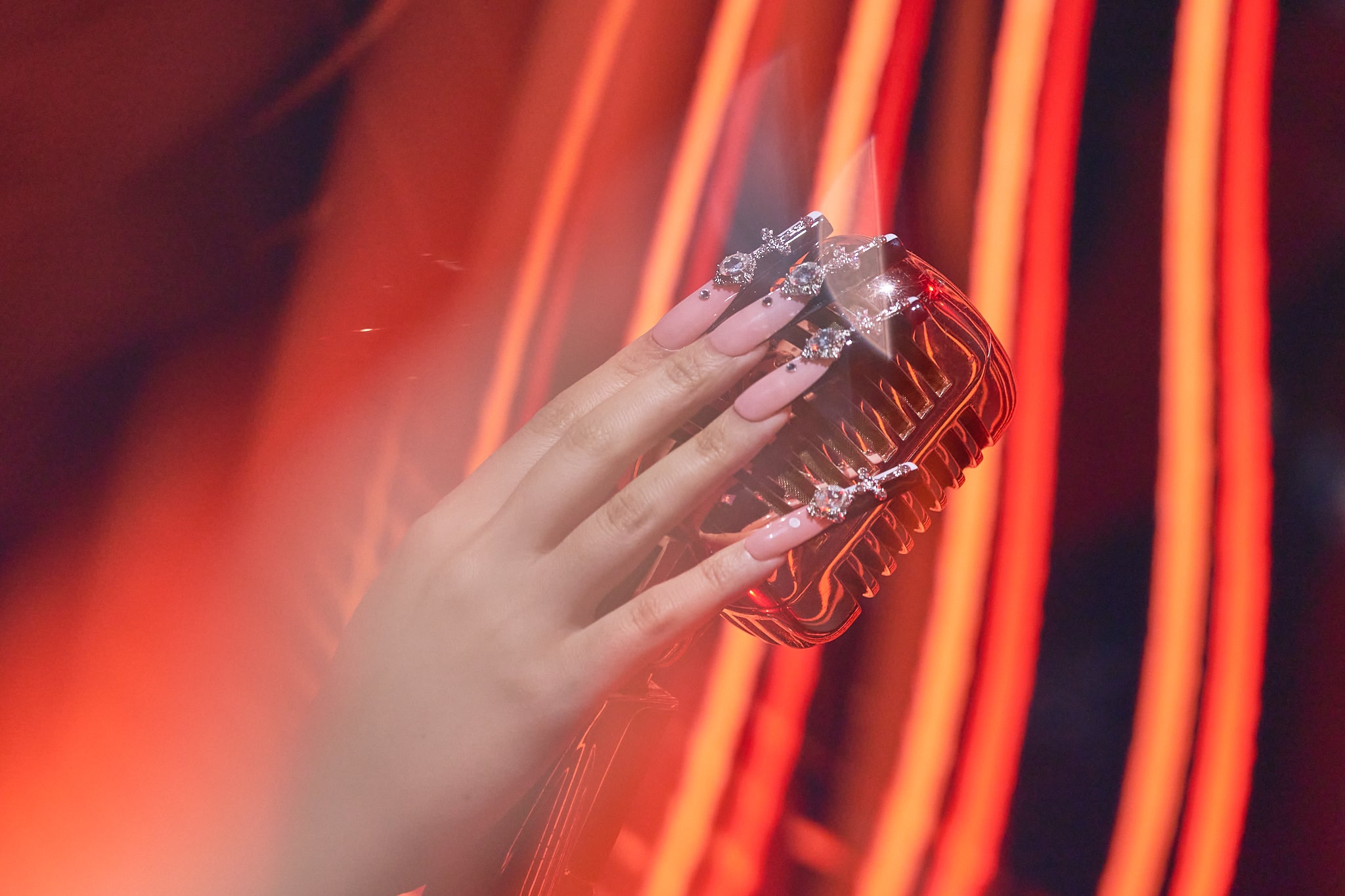 Hand with MiuNail Divine Plea press-on nails holding a silver microphone under vibrant orange stage lighting.