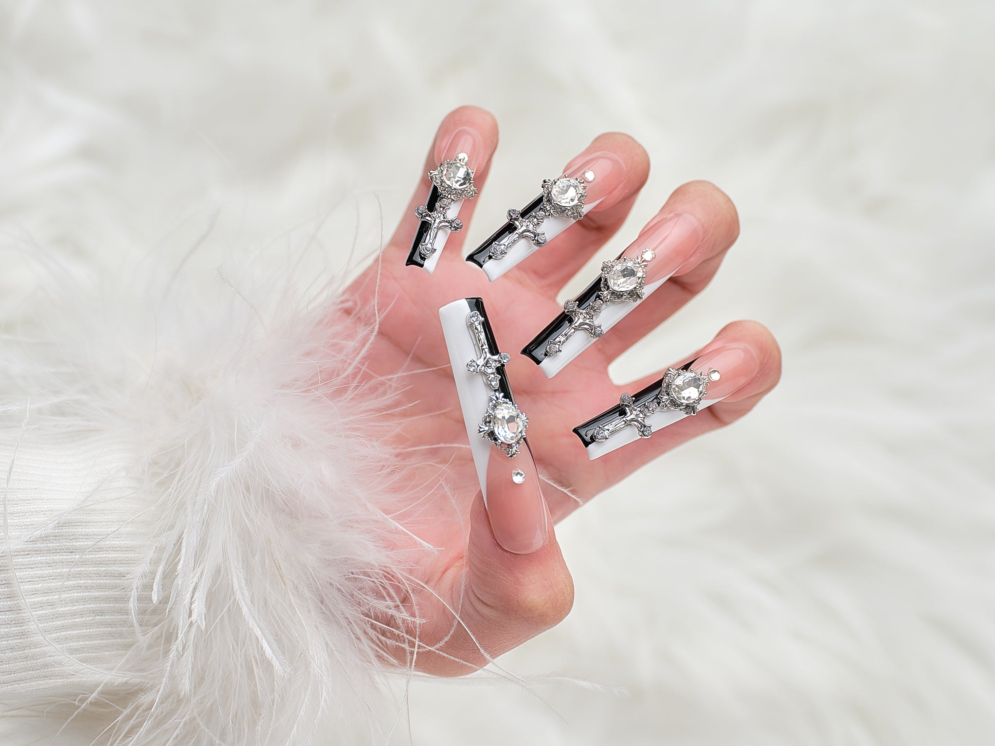 Close-up of a hand wearing MiuNail Divine Plea press-on nails, featuring rhinestones and silver cross charms over a nude, black, and white design, against a fluffy white background.